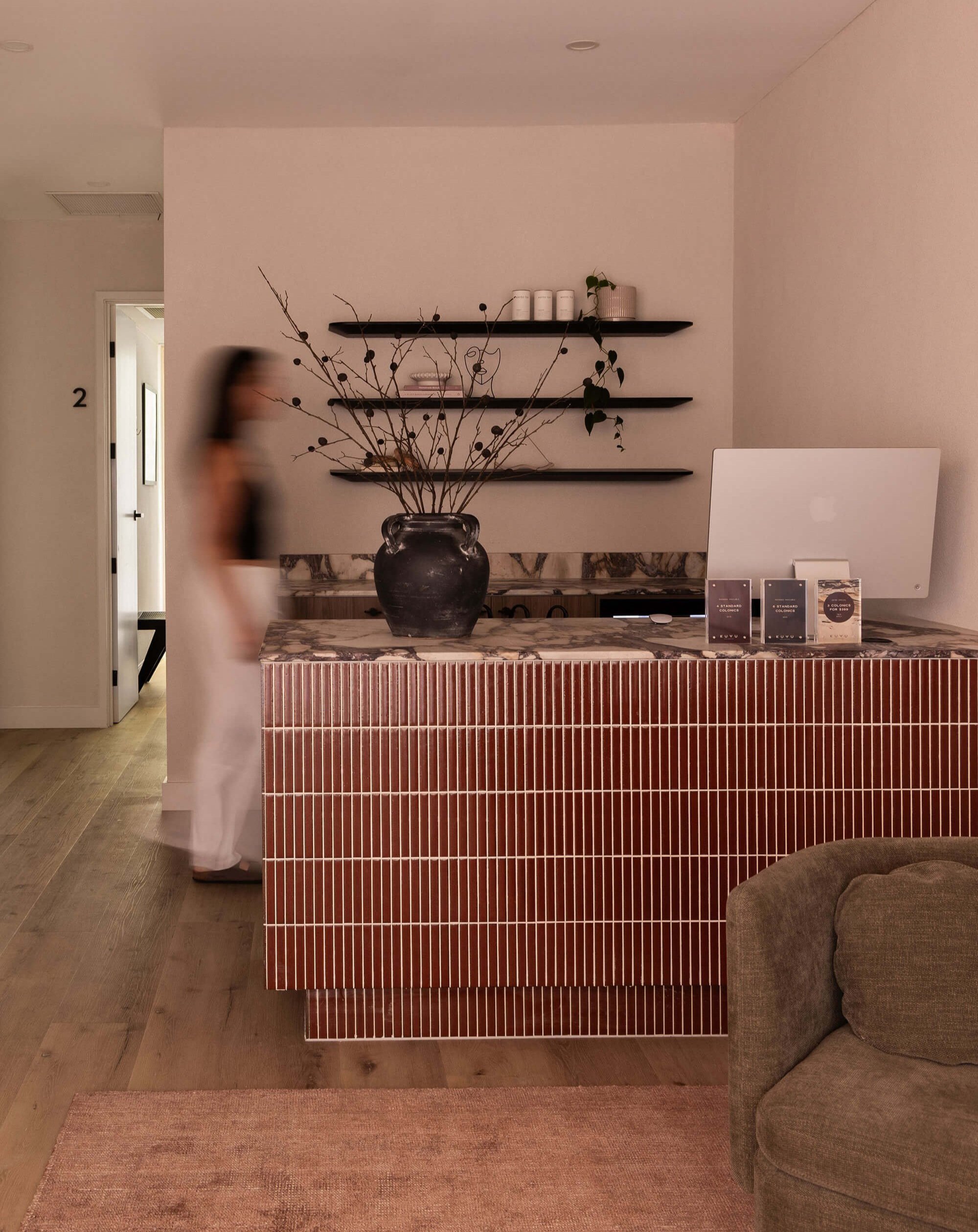 Modern reception area with marbled counter, large black vase with branches, computer, and wall shelves with decor