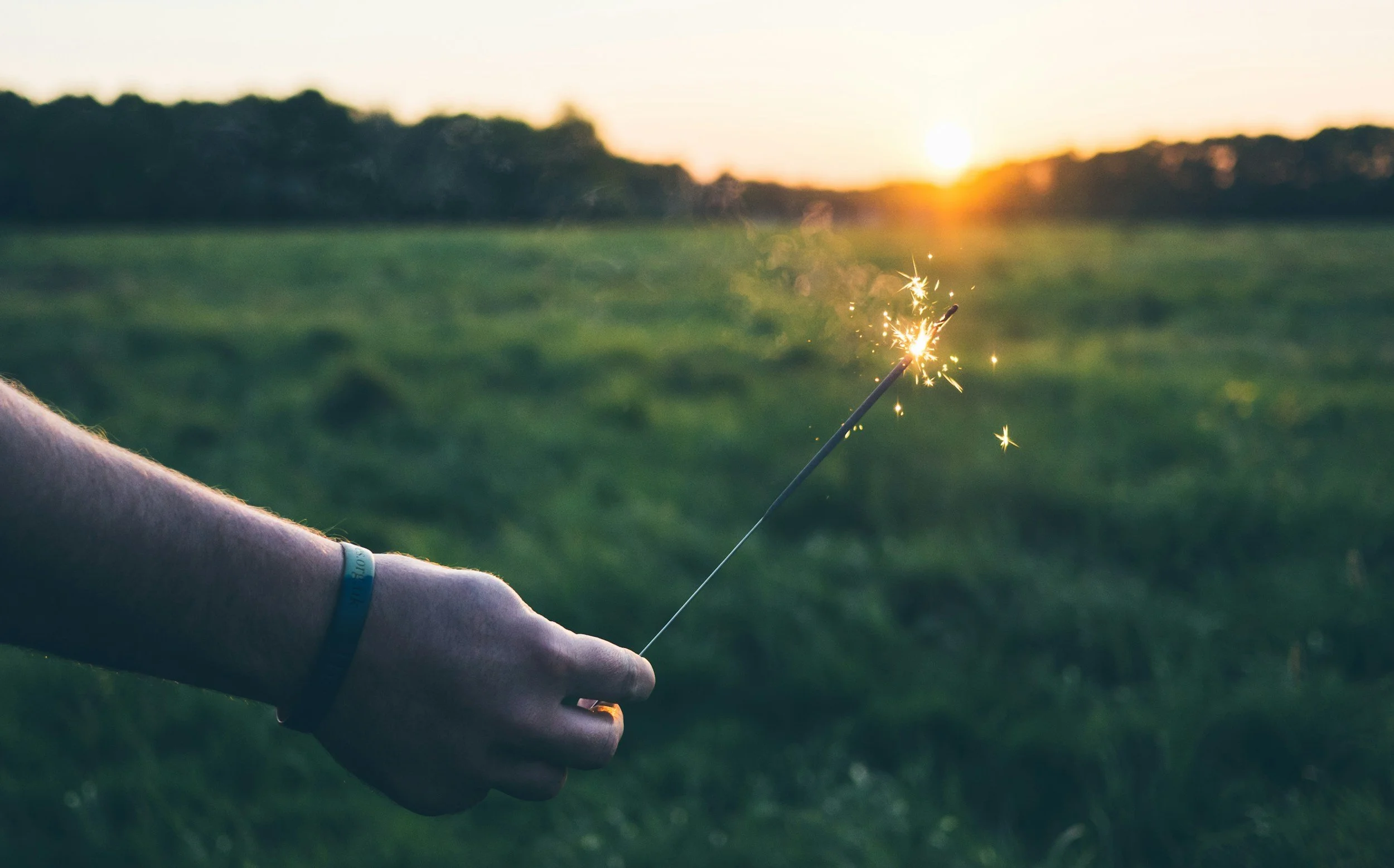 Person holding a lit sparkler outdoors during sunset, with a green field and trees in the background.