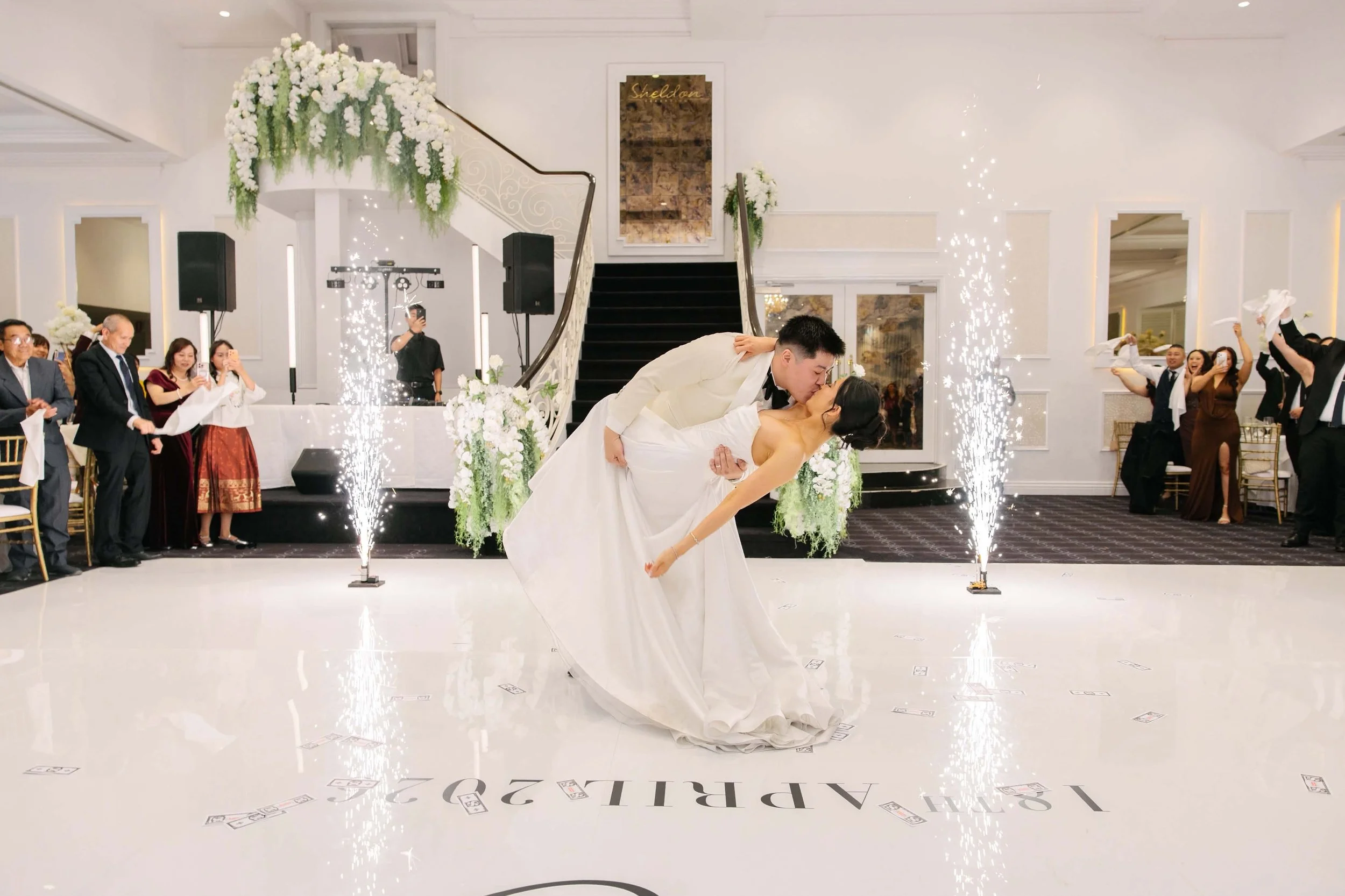 A bride and groom share a kiss during their wedding reception, with sparklers and guests celebrating around them in an elegant, decorated ballroom.