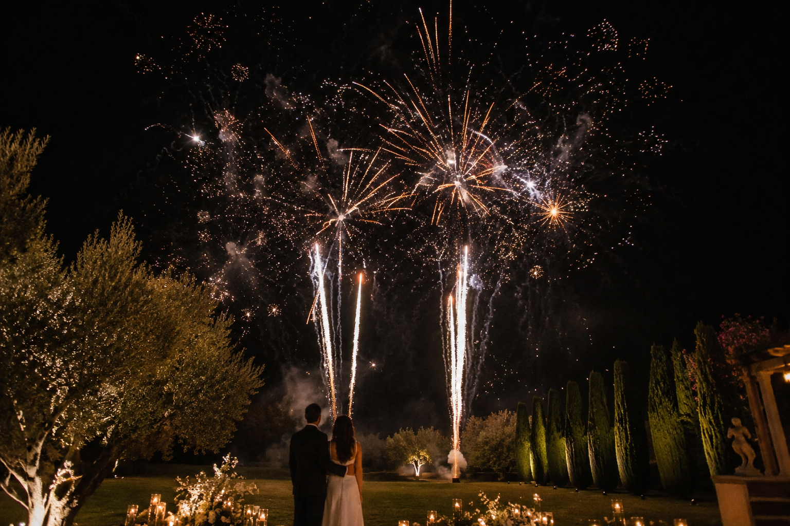 A couple in formal attire watching fireworks explode in the night sky, surrounded by lit candles and trees.