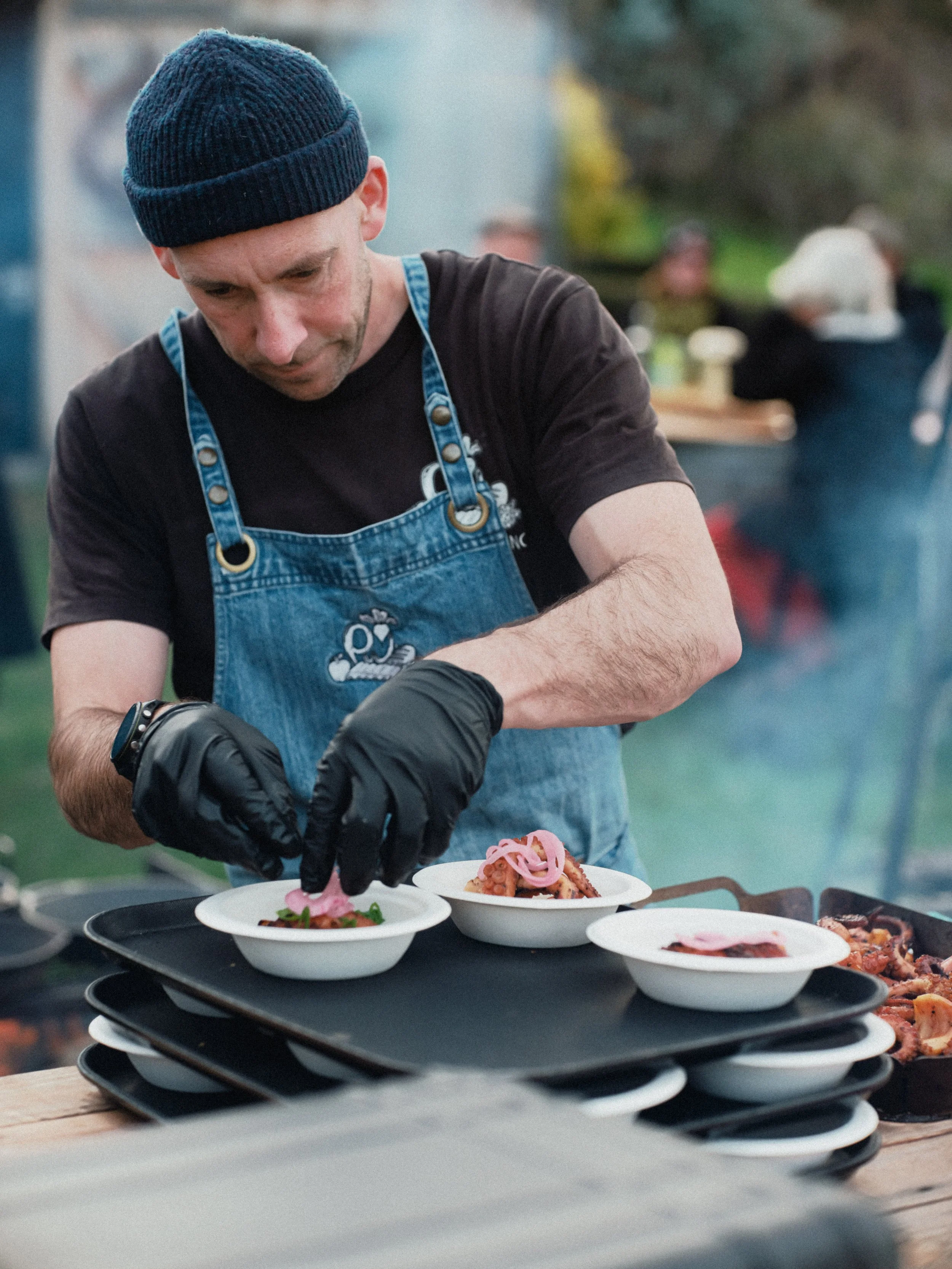 A man wearing a black shirt, blue apron, and navy beanie is preparing dishes at a food service event. Several plates with colorful food are stacked on a black tray in front of him, with additional food on the table.