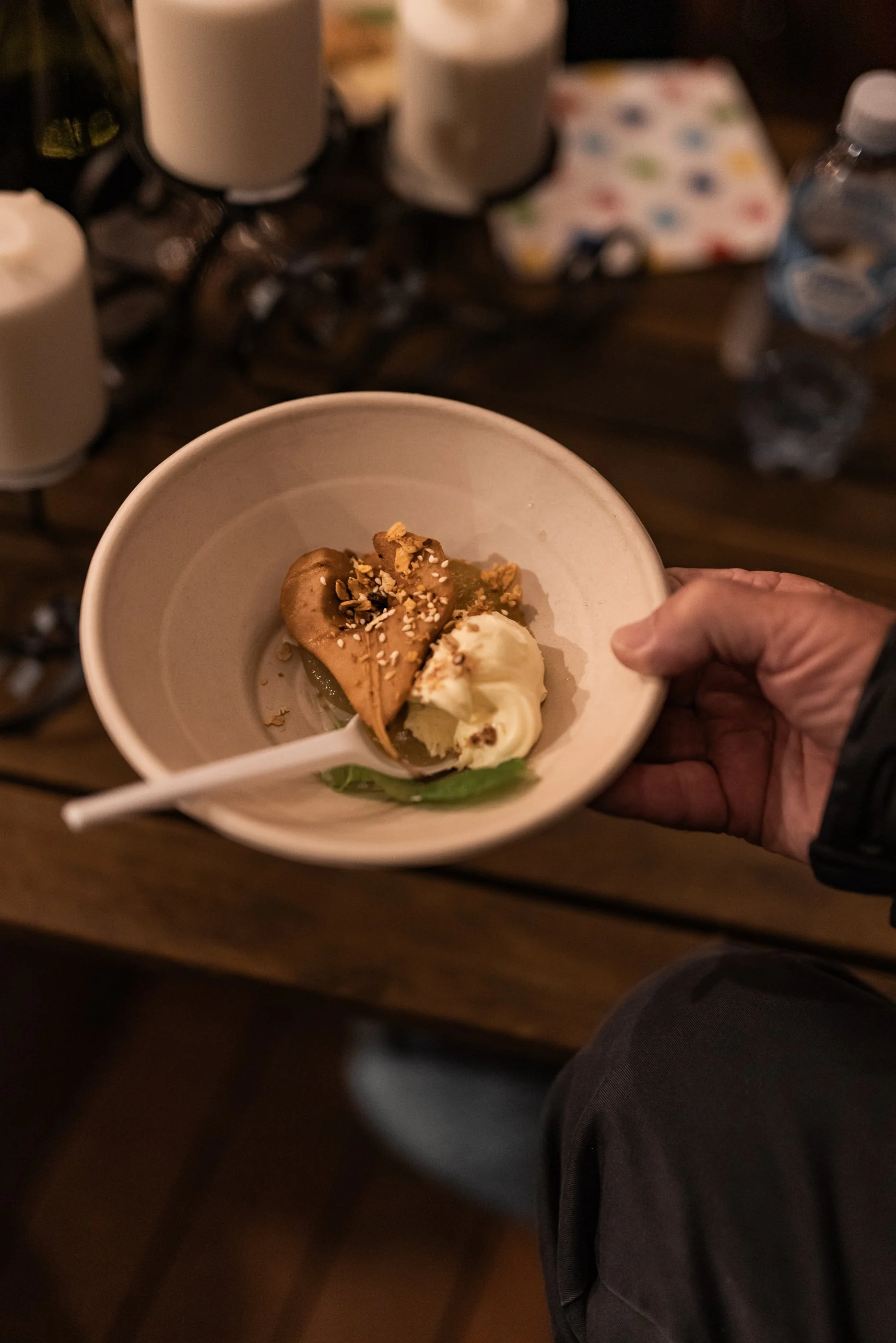 A person holding a bowl with a dessert that includes a poached pears, custard, and toppings like chopped nuts, with a wooden table and candles in the background.