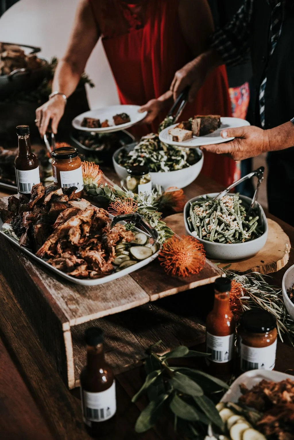 People serving and placing food on a table at a buffet-style meal with various dishes including salad, roasted meat, pasta salad, and condiments.