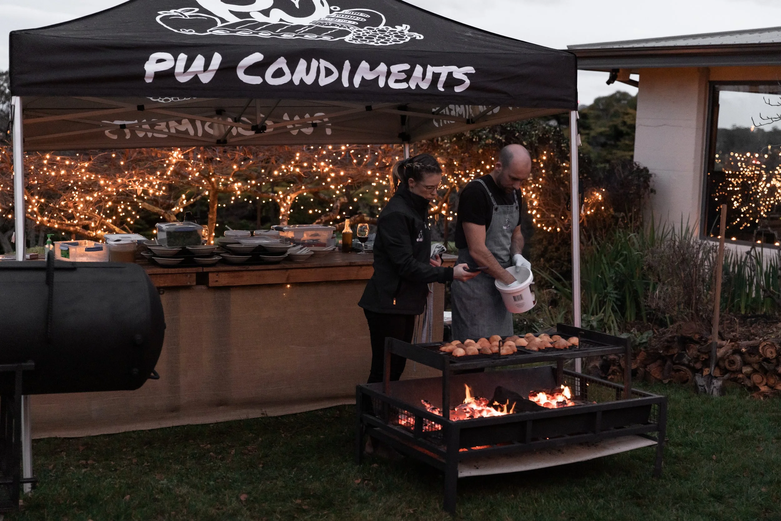 Two people grilling Octopus over fire at an outdoor food stand with a sign that reads "PW Condiments" under a canopy, with twinkling string lights in the background during dusk.