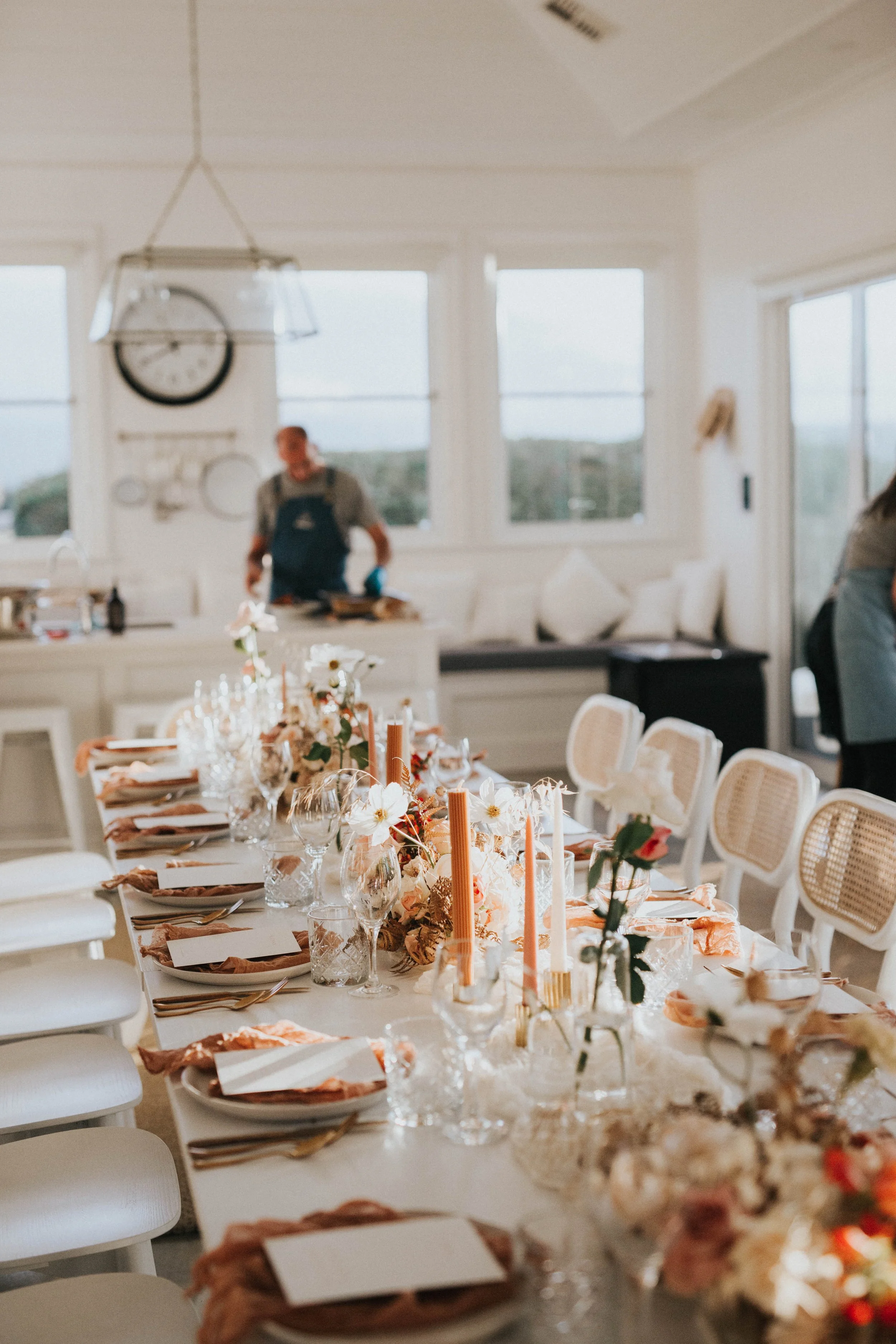 A festively decorated dining table with floral centerpieces, candles, glasses, and plates set for a meal, in a bright room with large windows and the chef standing in the background wearing an apron.