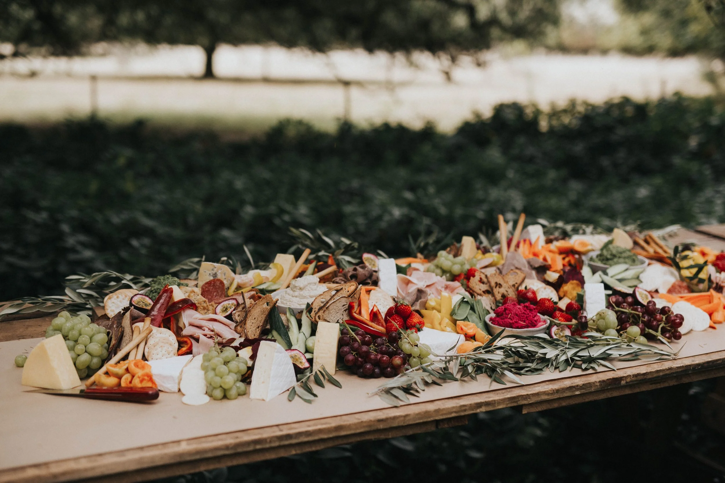 A large cheese and charcuterie platter with grapes, strawberries, crackers, and various cheeses on a wooden board outdoors.