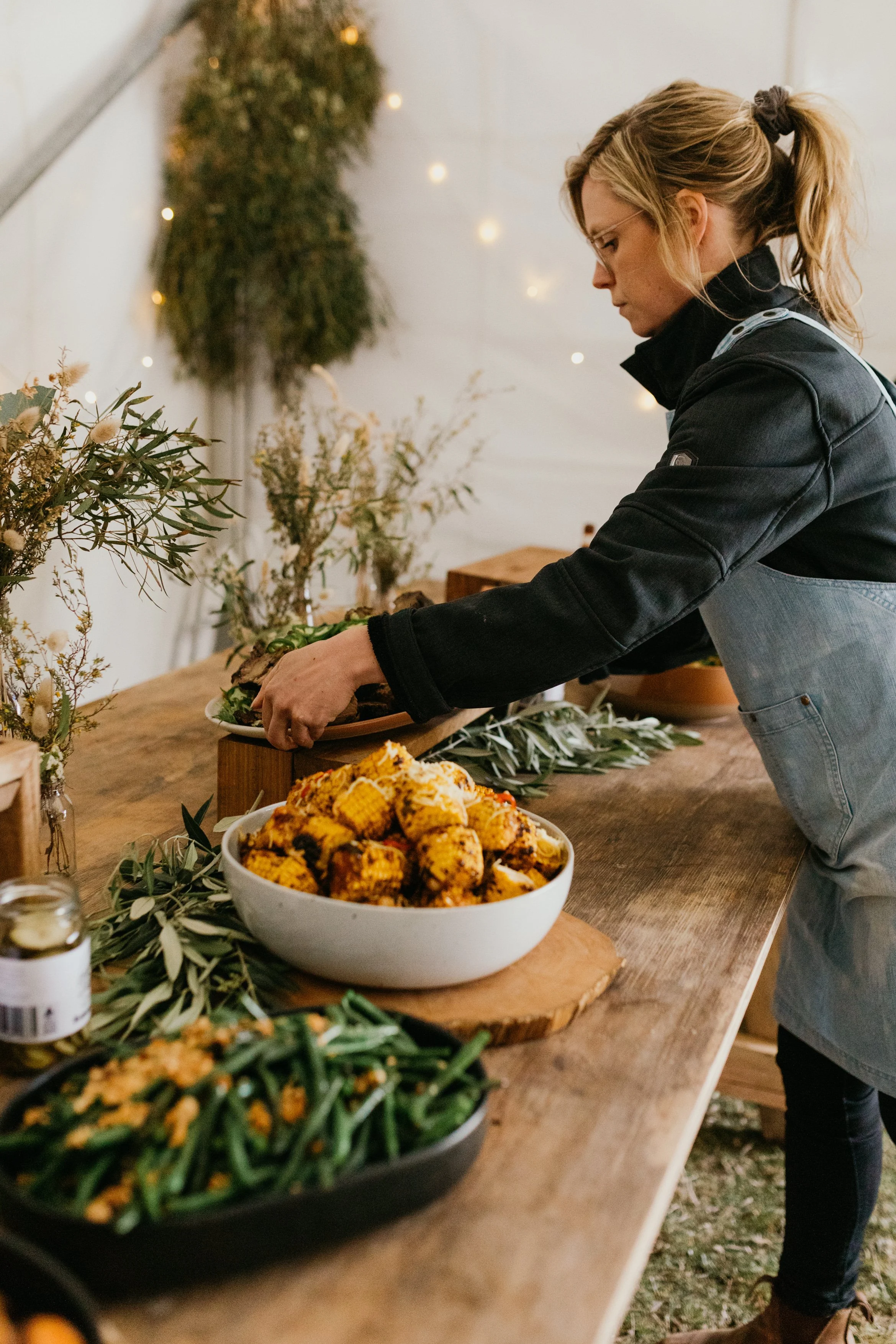 A woman arranging a platter of food at a table with festive decorations, including greenery and fairy lights in the background.