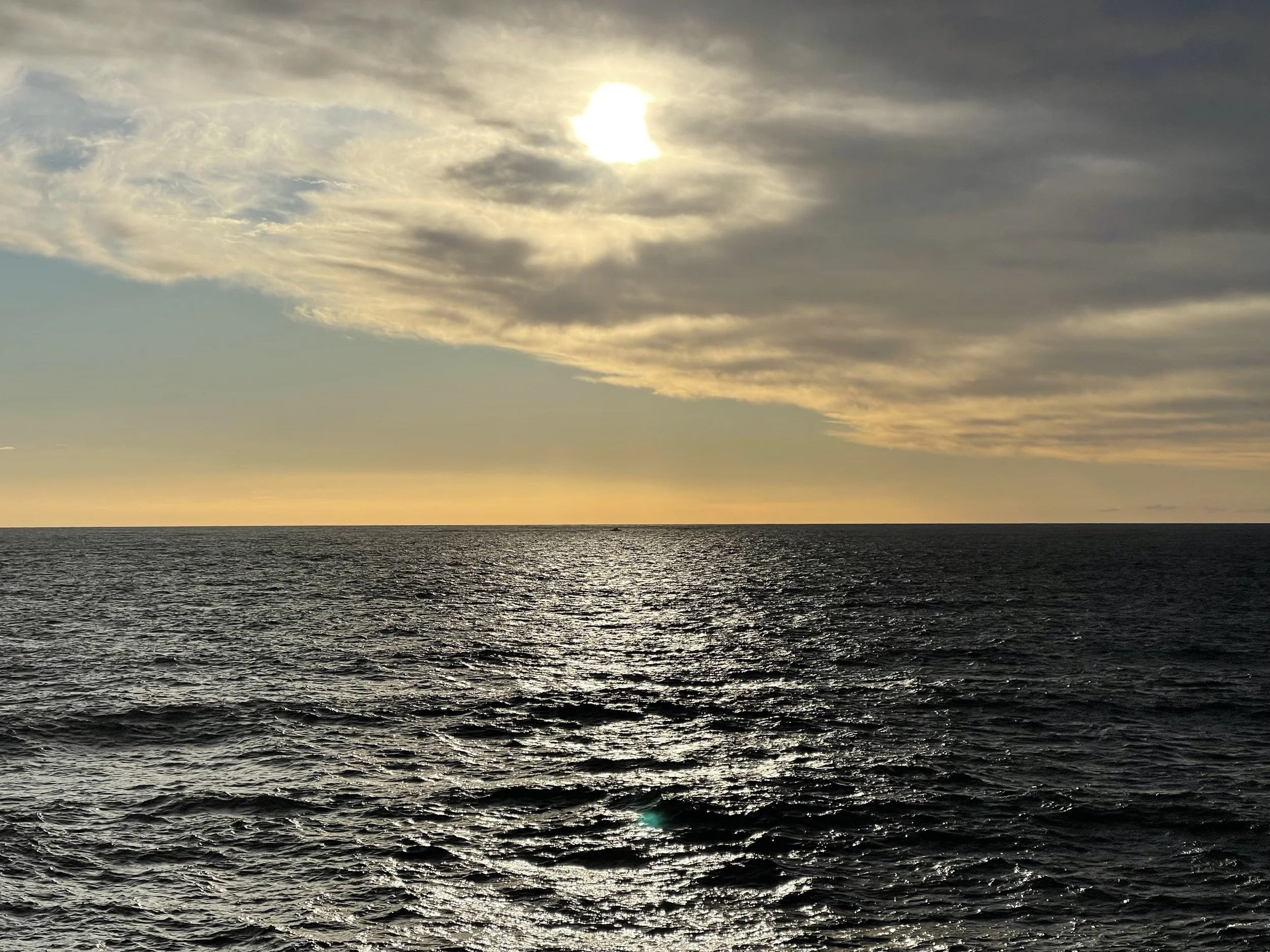 The image shows a sunset over the ocean with a partly cloudy sky, the sun is partially covered by clouds, and the water surface appears textured with gentle waves.