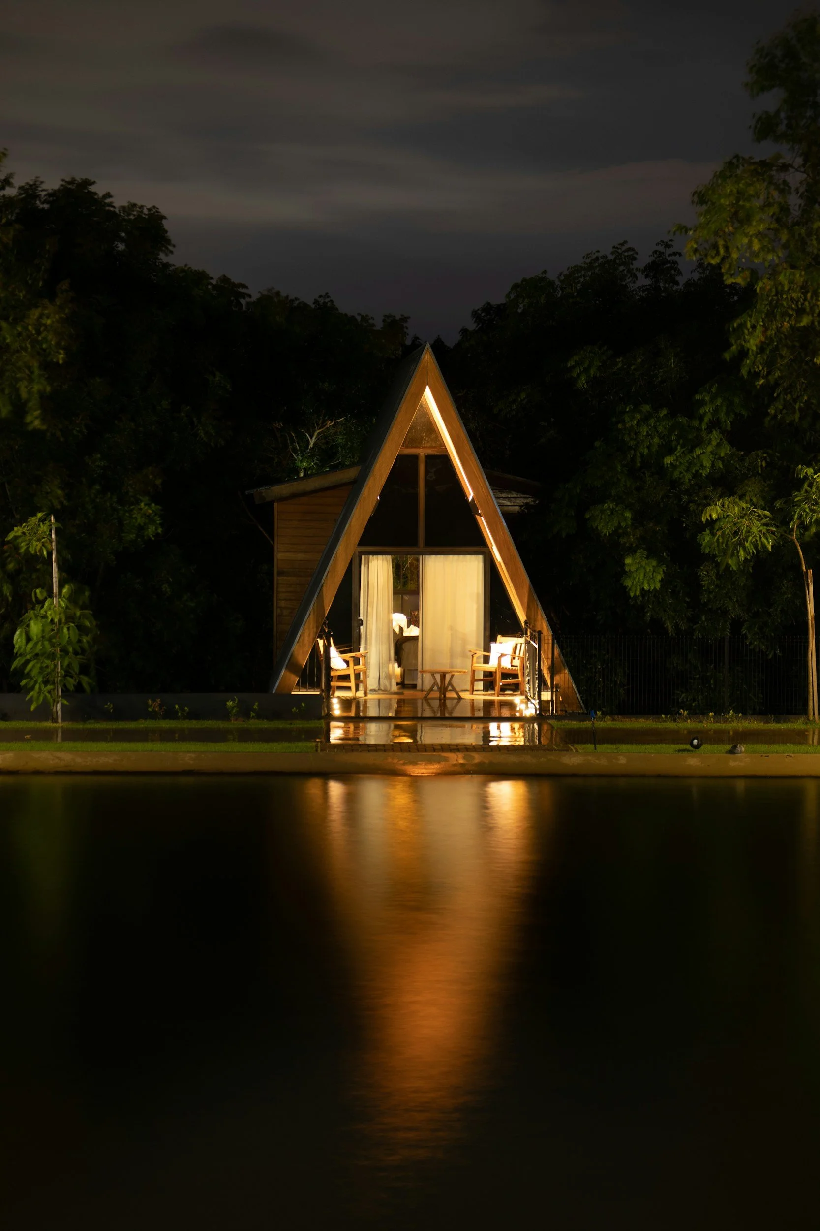 Aerial night view of a modern, A-frame cabin on a lake, illuminated from within, surrounded by trees, with calm water reflecting the building and dark cloudy sky.