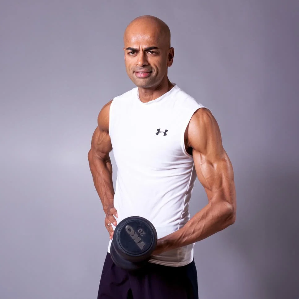 A muscular man with a shaved head lifting a dumbbell, wearing a white sleeveless athletic shirt and black shorts against a plain gray background.