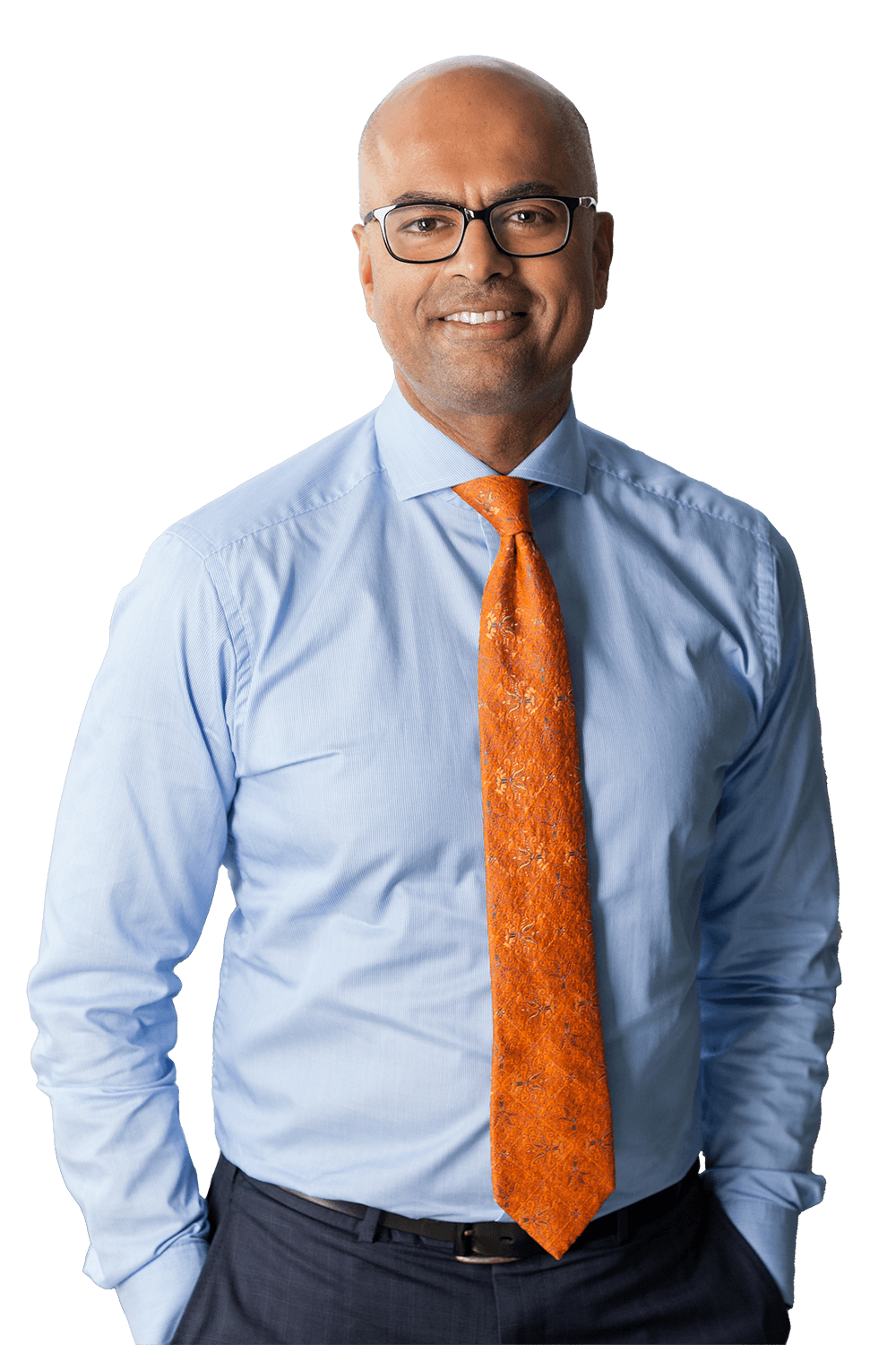 A man wearing glasses, a light blue dress shirt, and an orange patterned tie, smiling and standing with his hands in his pockets.