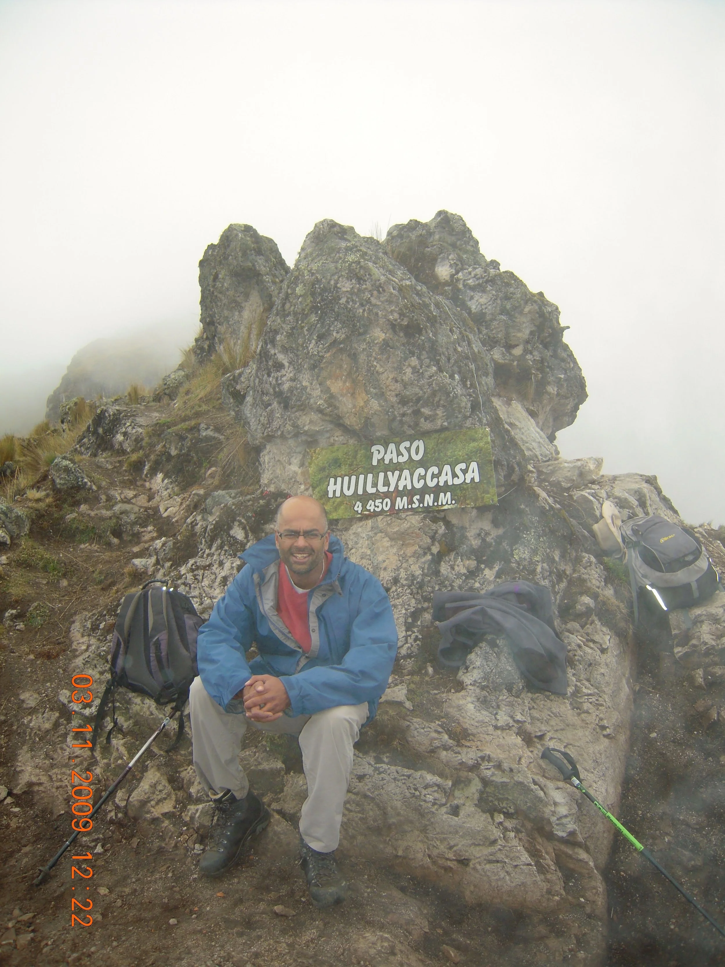 A man wearing glasses and a blue jacket sitting on rocks at the summit of Huilluyaccasa mountain, elevation 4,450 meters above sea level, with a foggy background and a sign indicating the mountain's name and elevation.