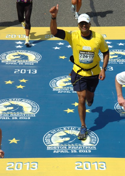 Male marathon runner crossing finish line at the Boston Marathon on April 15, 2013, wearing a yellow shirt, black shorts, and a white cap, with a race bib number 22619, raising his right fist in celebration.