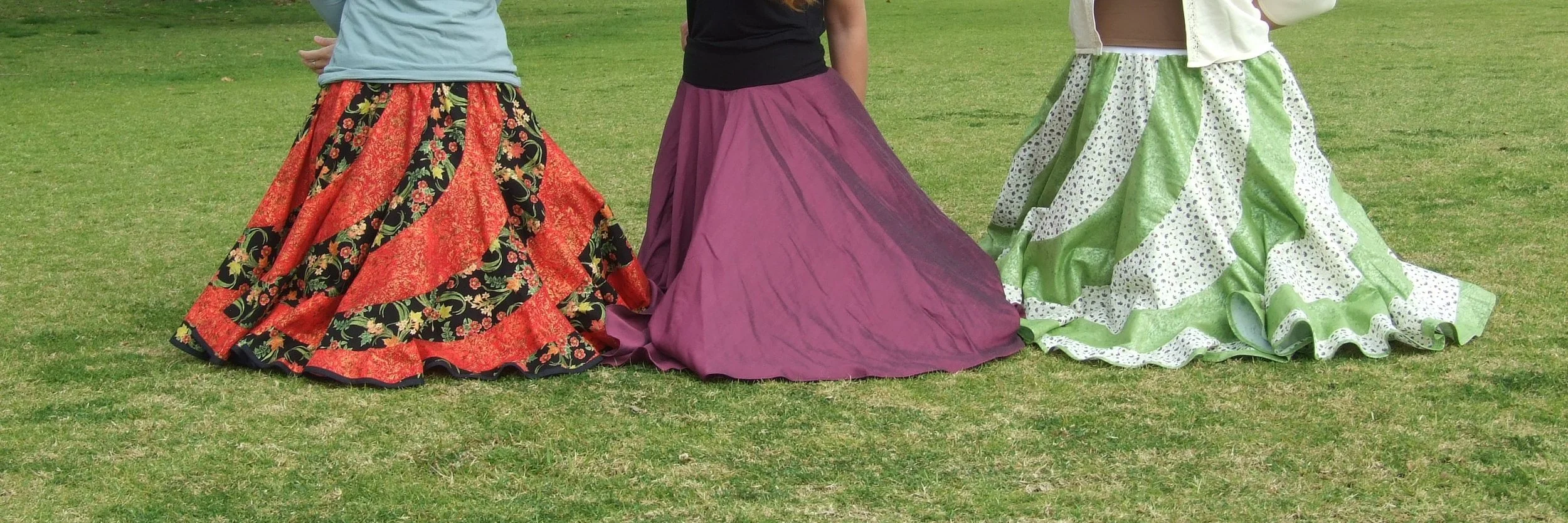 Three women kneeling on grass wearing Designs by Uriel skirts: a red, gold and black Signature Swirl Skirt, a Midnight Rose Classic Circle Skirt, and a green, cream, purple and turquoise Signature Swirl Skirt with pearlised gold detailing.