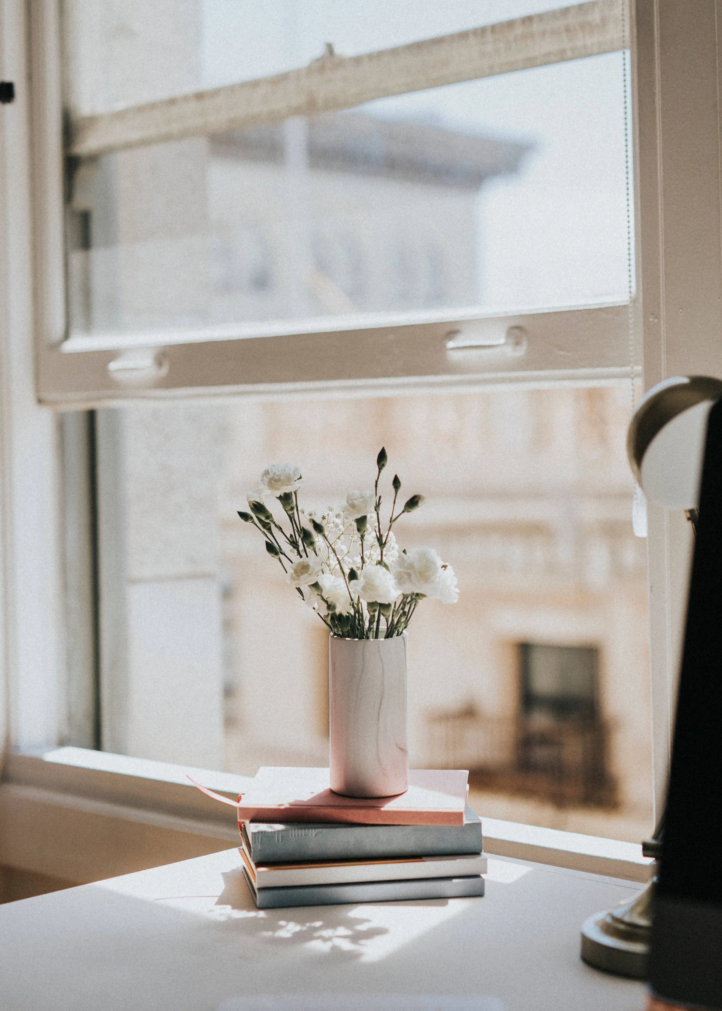 A stack of books on a table under a window, with a pinkish vase containing white flowers, and a lamp to the right.
