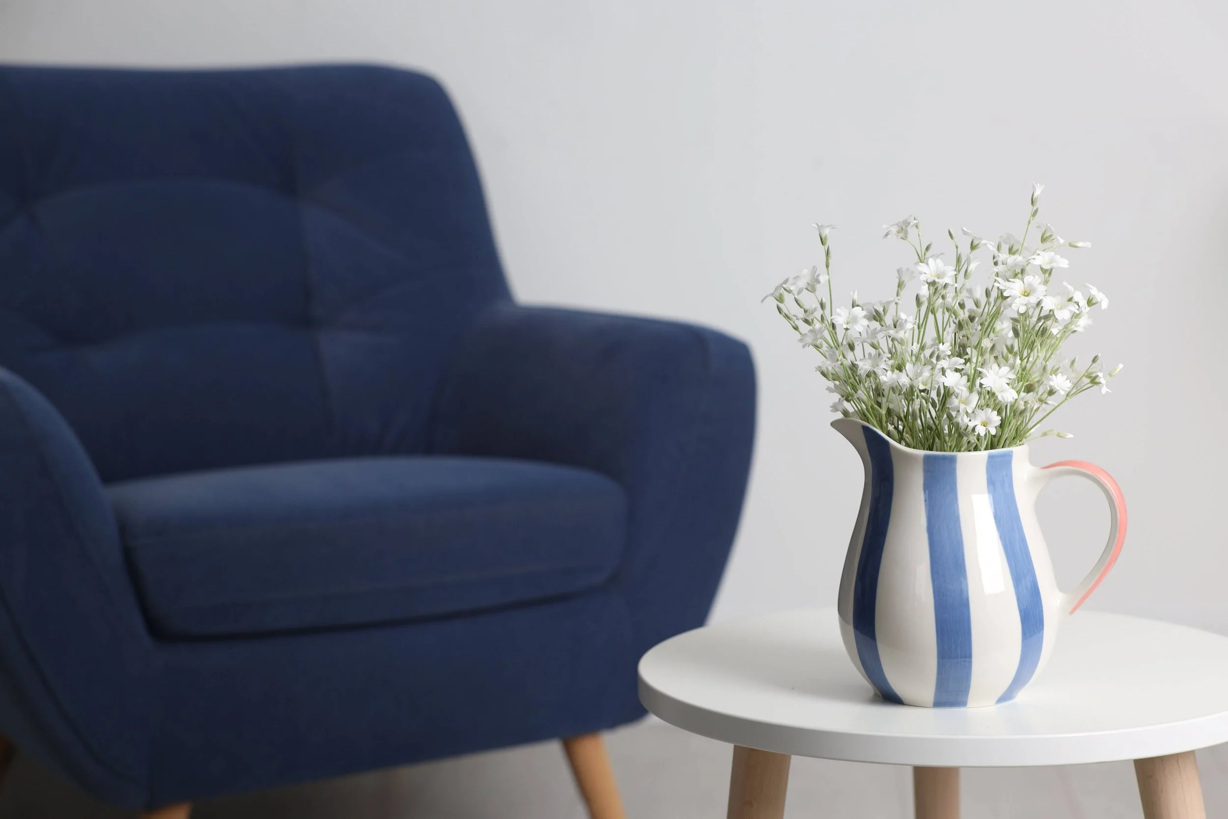 A white side table with light wooden legs holding a white vase with vertical blue stripes, filled with white flowers, next to a dark blue upholstered chair with button tufting.