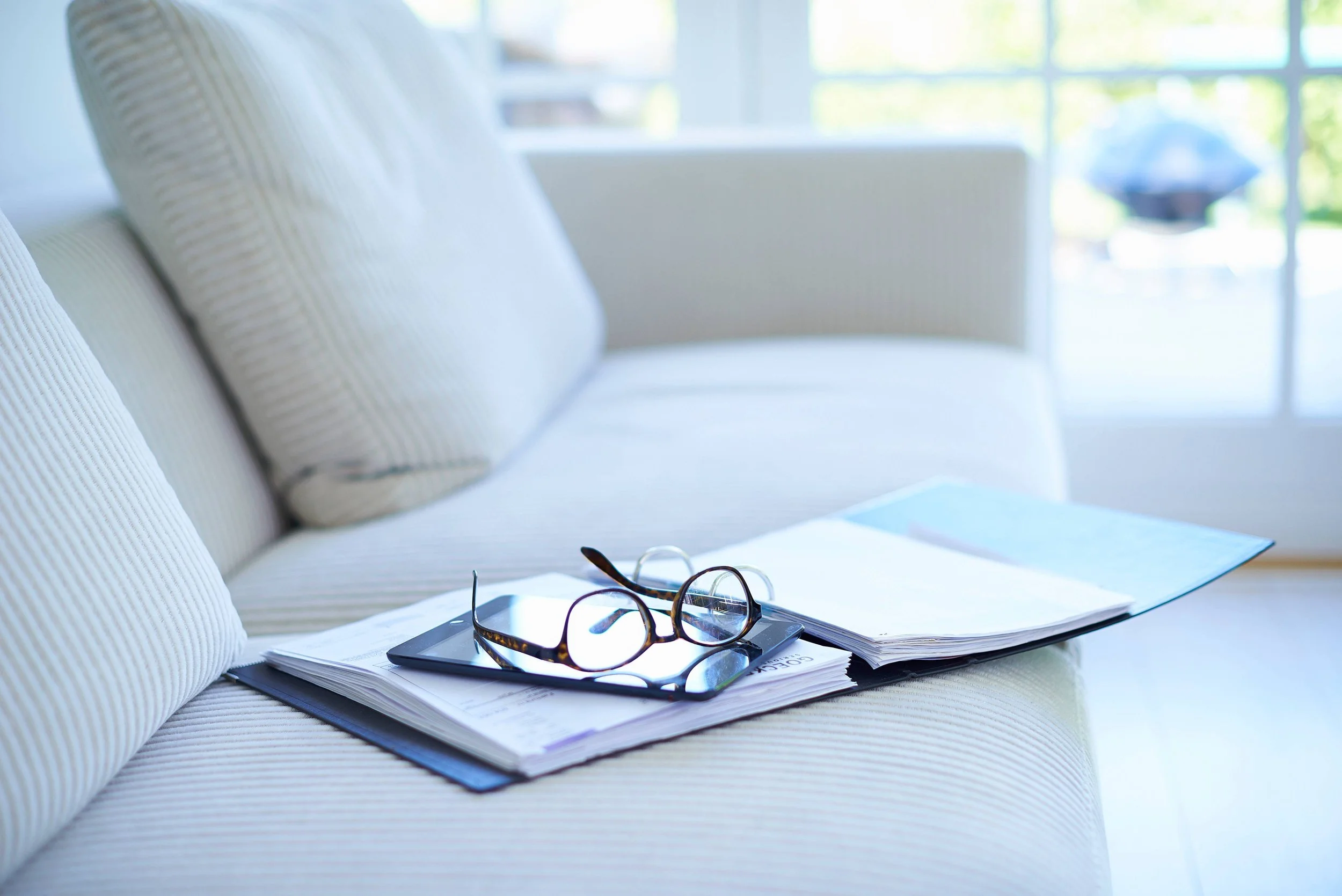A white couch with a notebook, a pair of eyeglasses, and a smartphone resting on it. The background features large windows with sunlight streaming in, creating a bright and airy atmosphere.