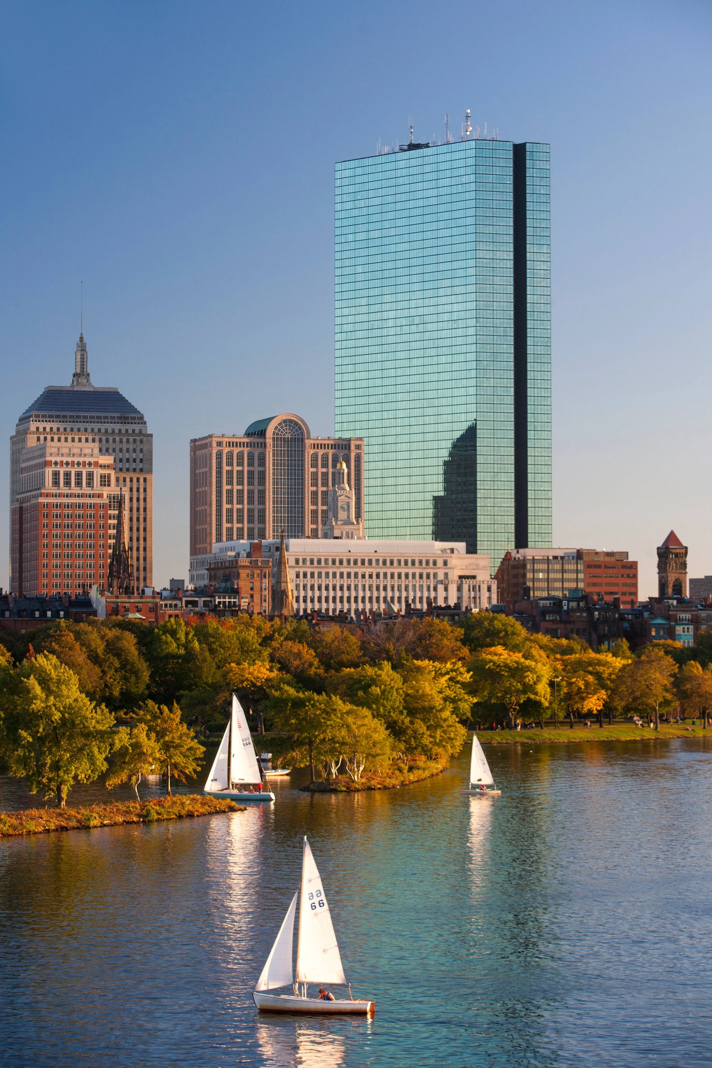 Sailboats on a river in front of a city skyline during sunset, with green trees along the riverbank and modern tall buildings in the background.