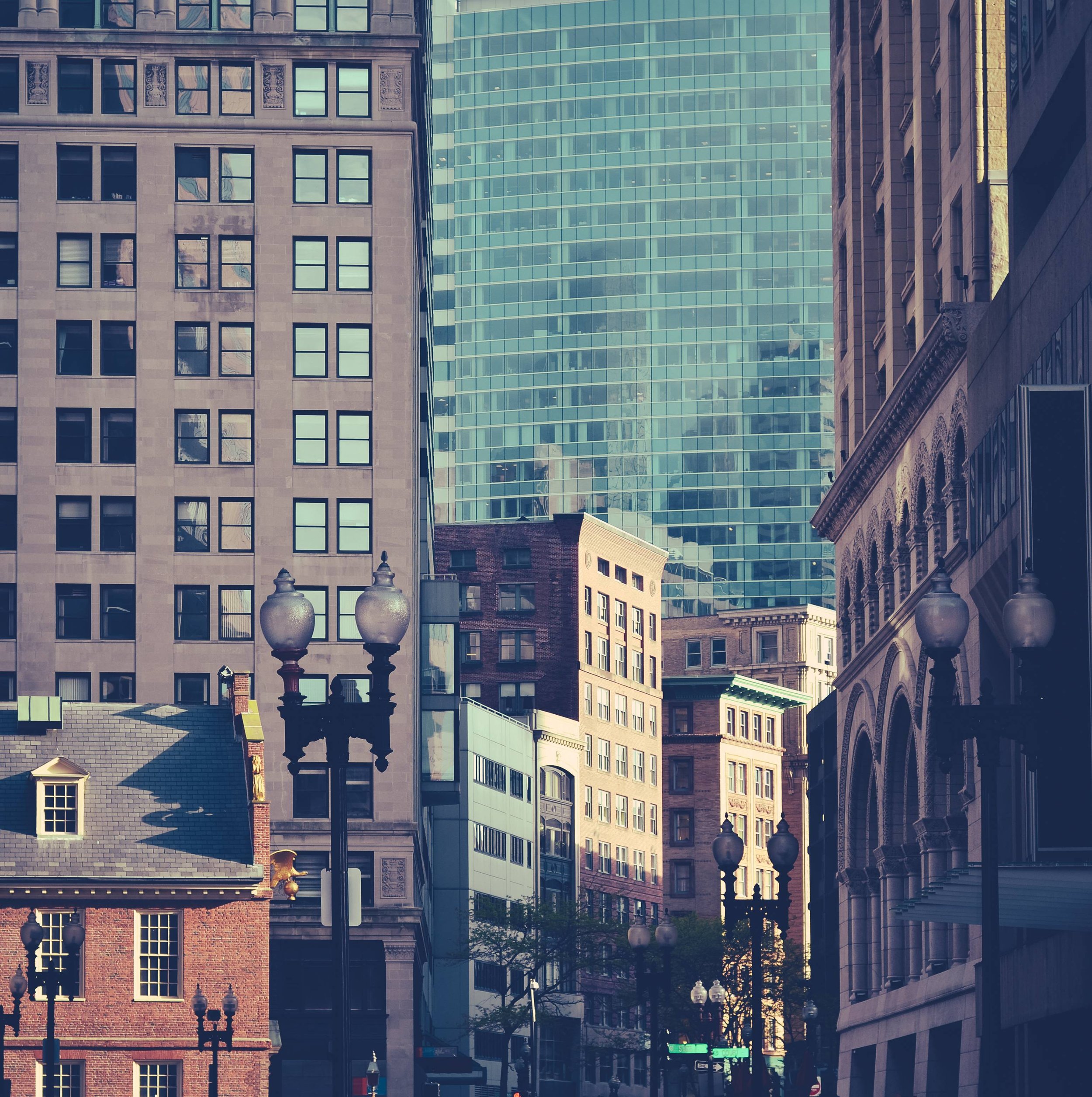 Tall city buildings with glass and brick facades in an urban downtown area, with street lamps and signs visible at street level.
