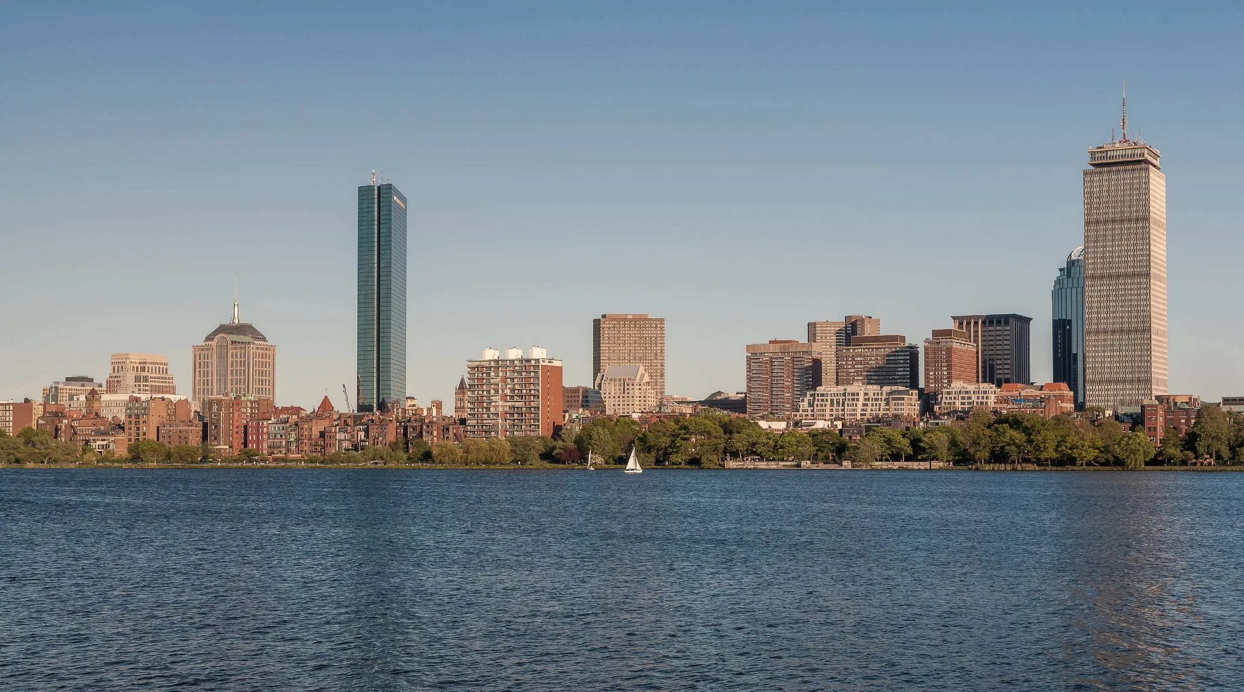 A city skyline with tall buildings, viewed across a body of water with small sailboats.