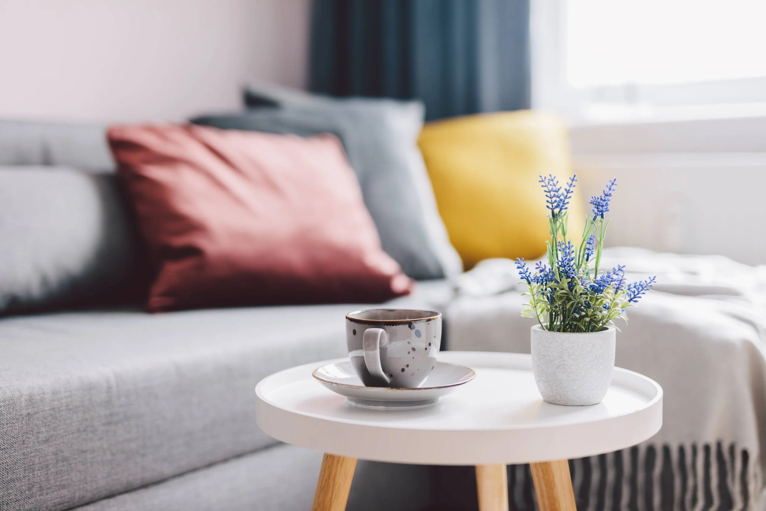 Living room scene with a gray sofa adorned with pink, gray, and yellow cushions, a small white side table holding a potted lavender plant and a polka-dotted coffee cup, and a window with blue curtains in the background.
