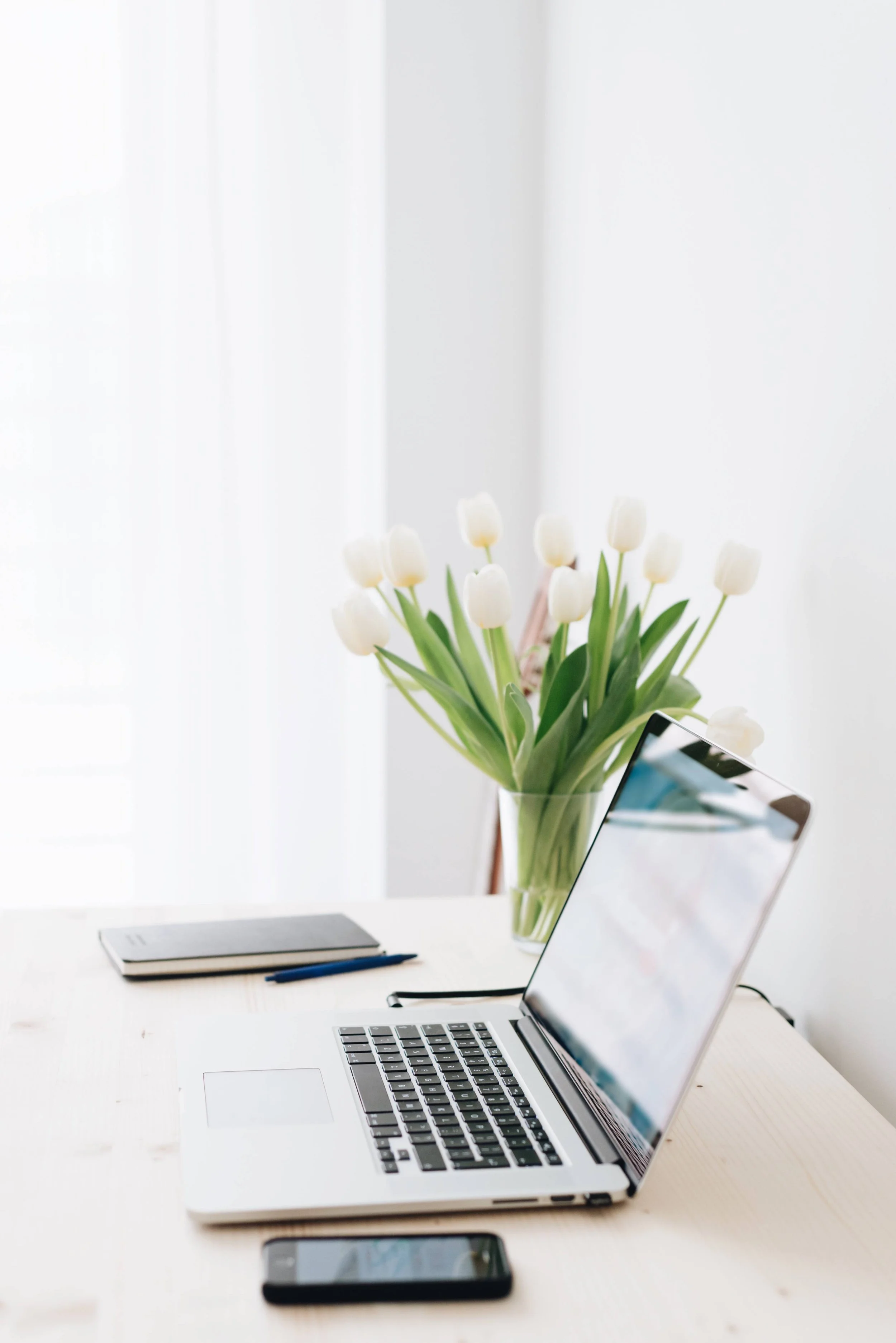 A workspace with a laptop, a smartphone, a notebook, pens, and a vase of white tulips on a white desk, with a bright window in the background.