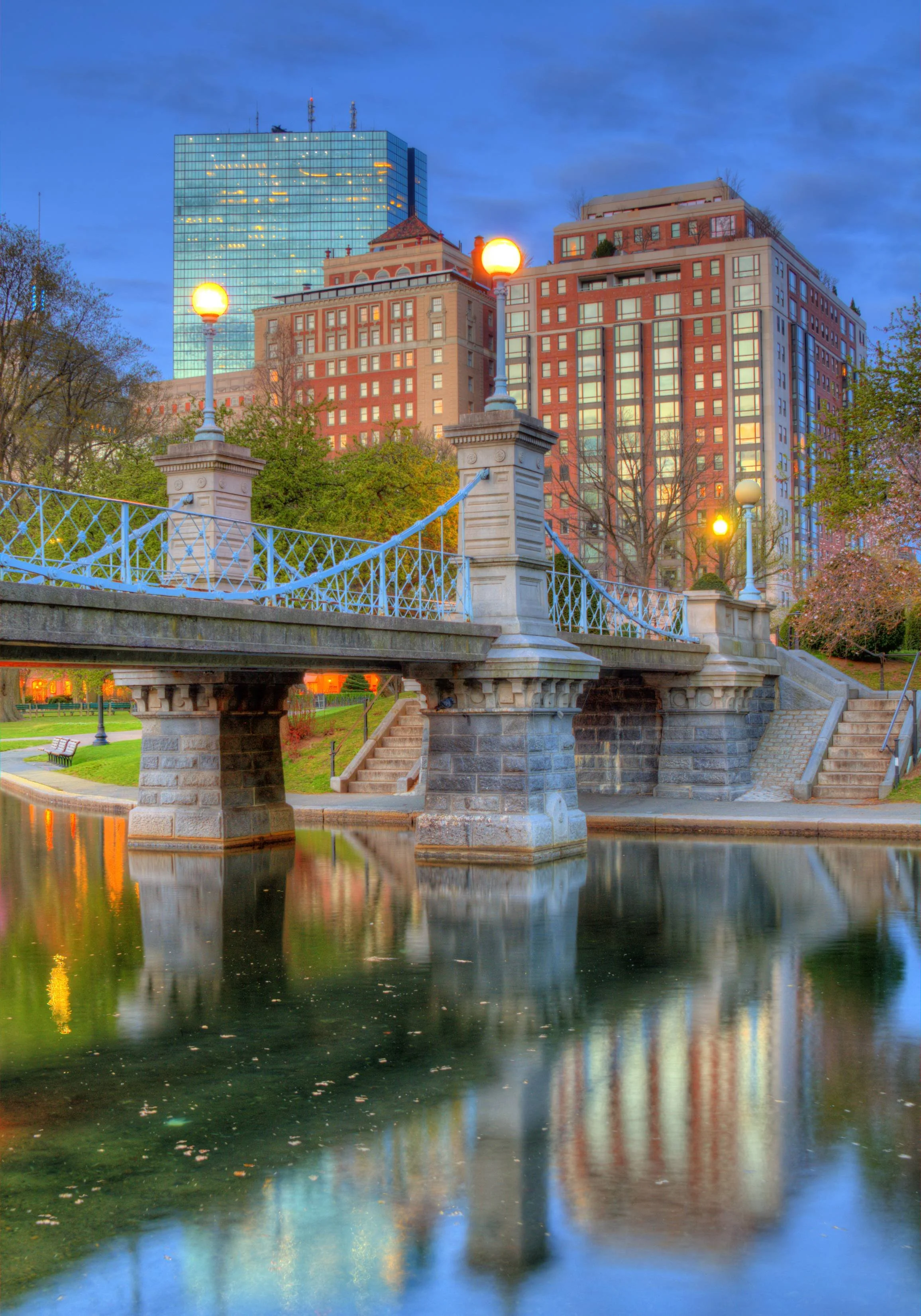 Cityscape at dusk with a bridge over a river, tall modern and historic buildings, trees, and street lamps reflecting on the water.