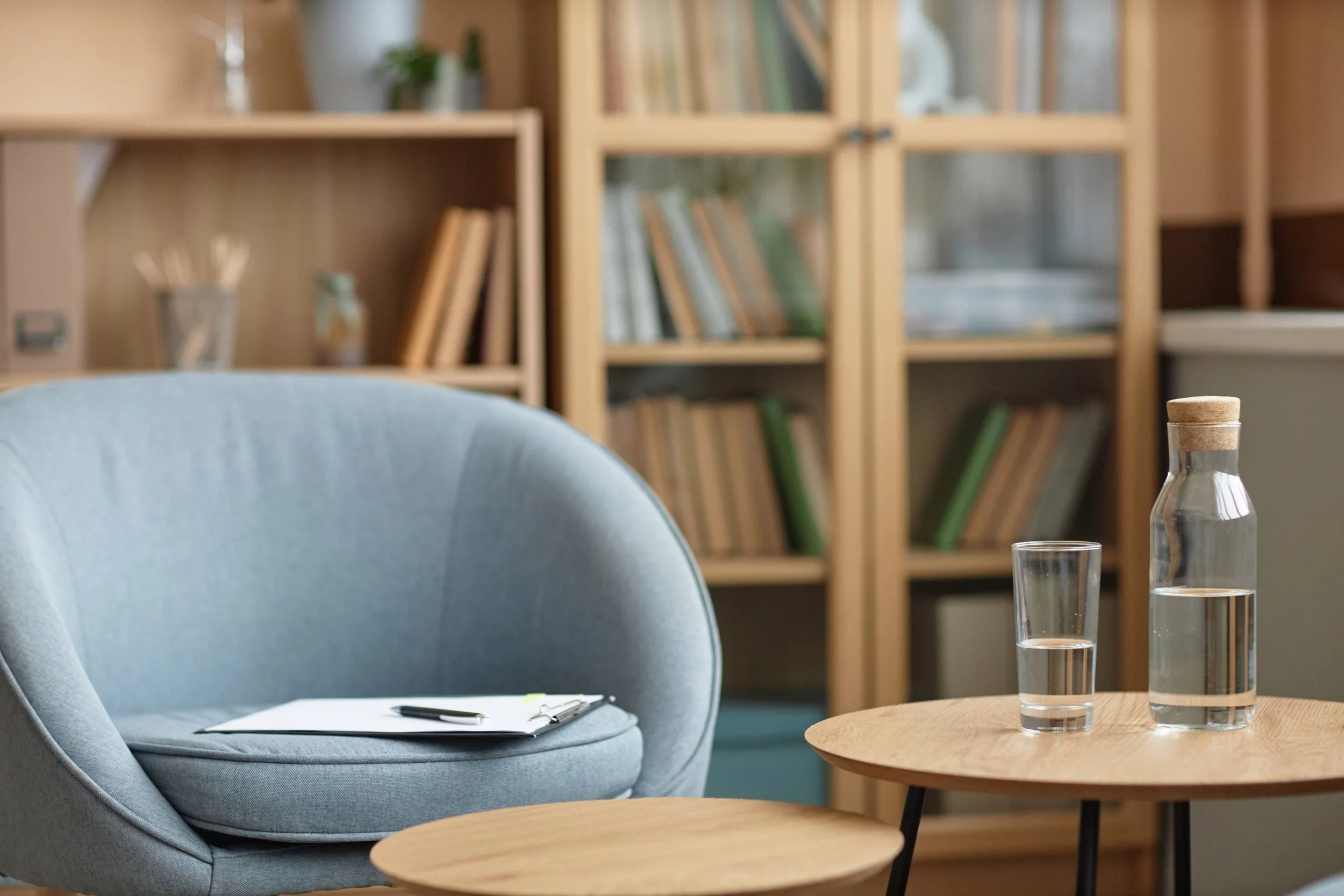 A cozy meeting room with a light blue armchair, a wooden table with glass water bottles, and a bookshelf filled with books in the background.