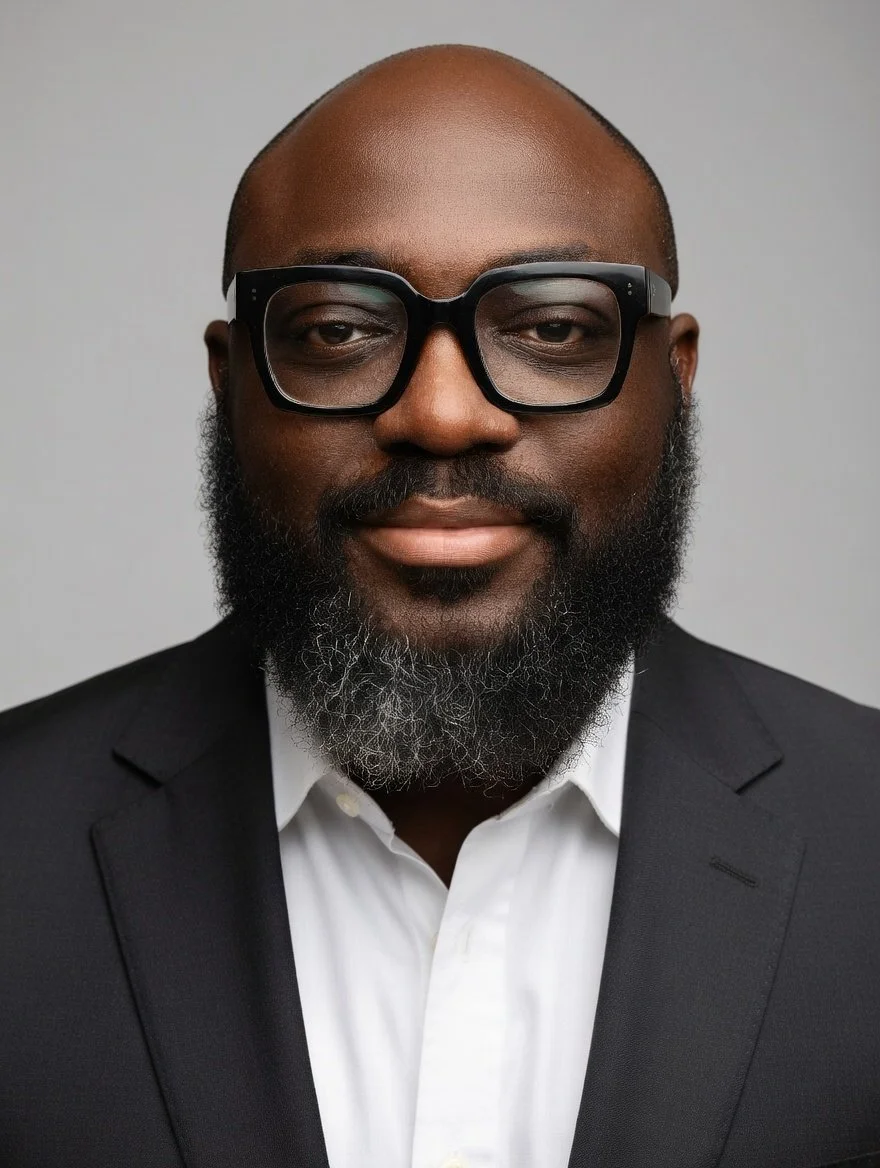 A portrait of a Black man with a beard and glasses, wearing a black suit jacket and white shirt, against a plain gray background.