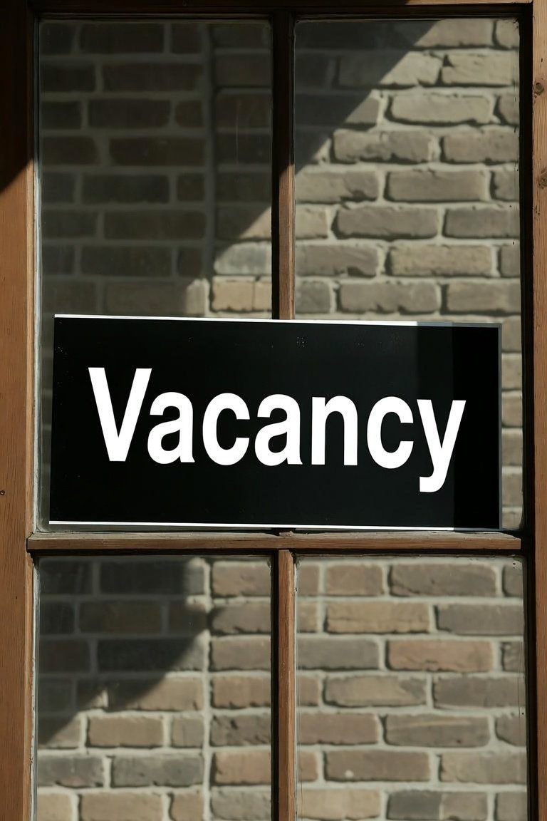 A window with wooden framing and a black sign that reads 'Vacancy' in bold white letters, with brick wall visible in the background.