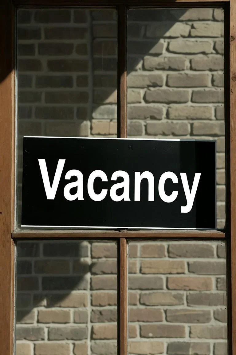 A window with wooden frame and glass panes, with a black sign that says "Vacancy" in white letters on it, hanging on the window.