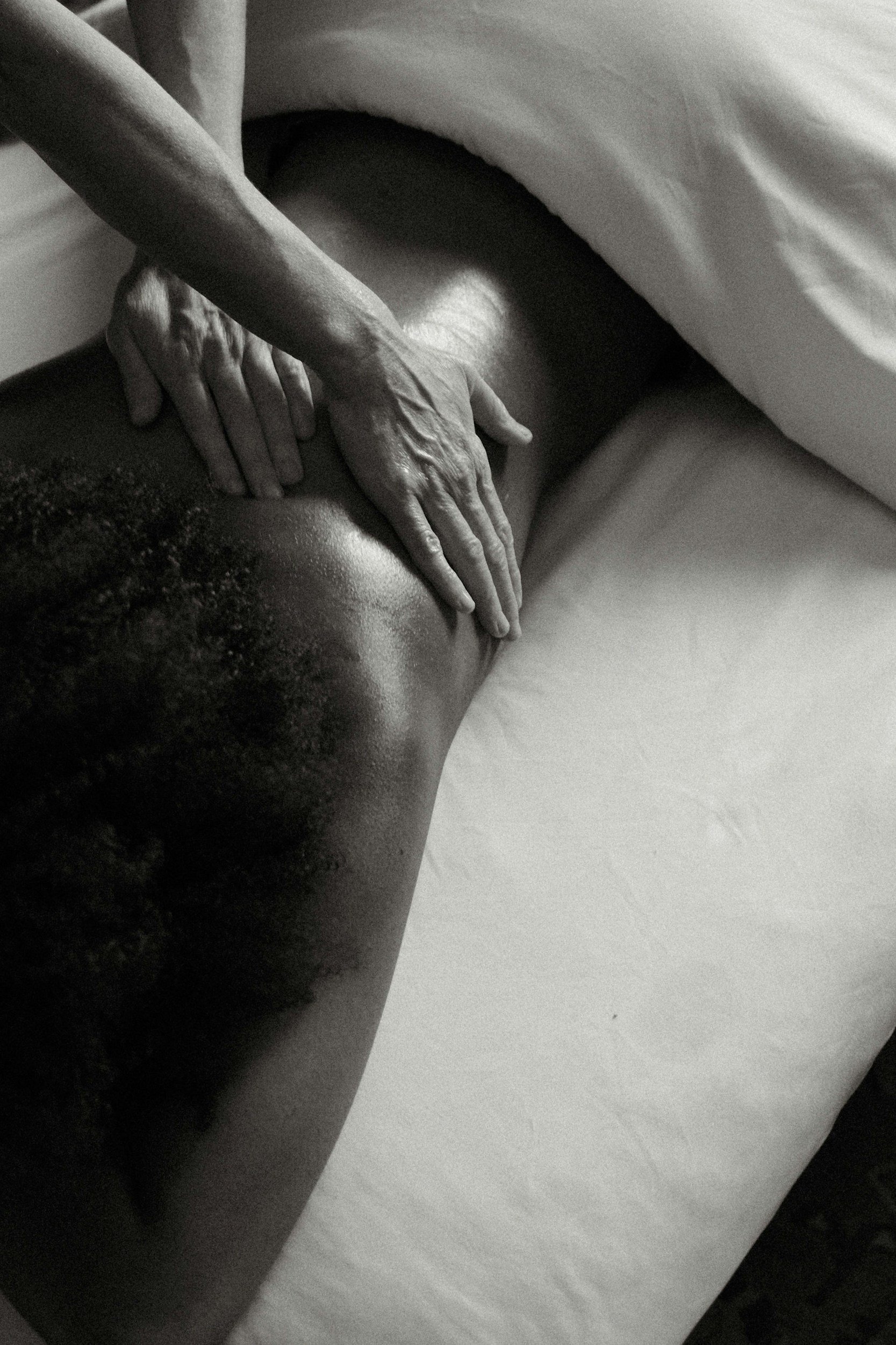 Close-up of a person's hands resting on their thighs, lying on a bed with white sheets, black-and-white photograph.