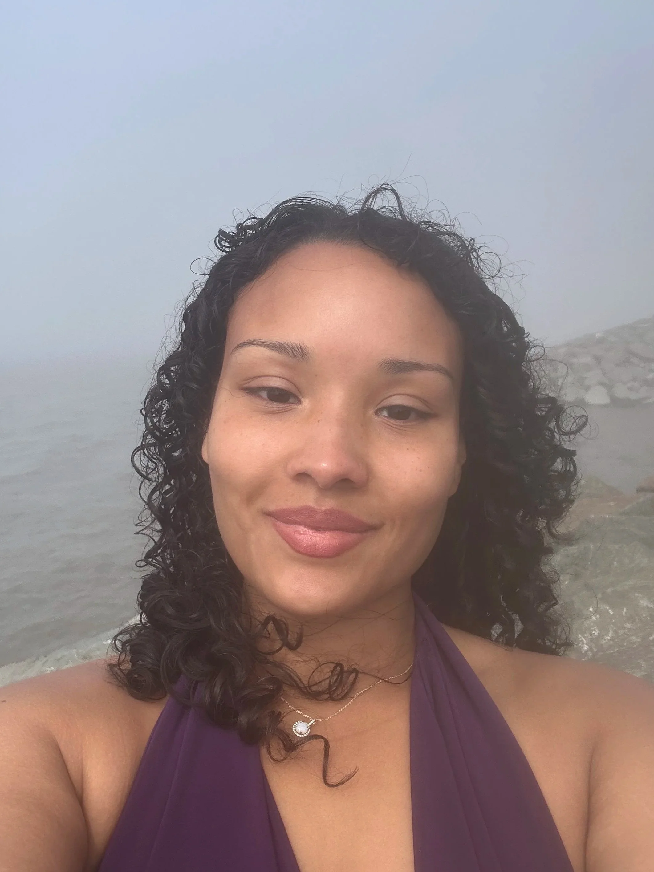 A woman with curly dark hair smiling at the beach with a cloudy sky and ocean in the background.