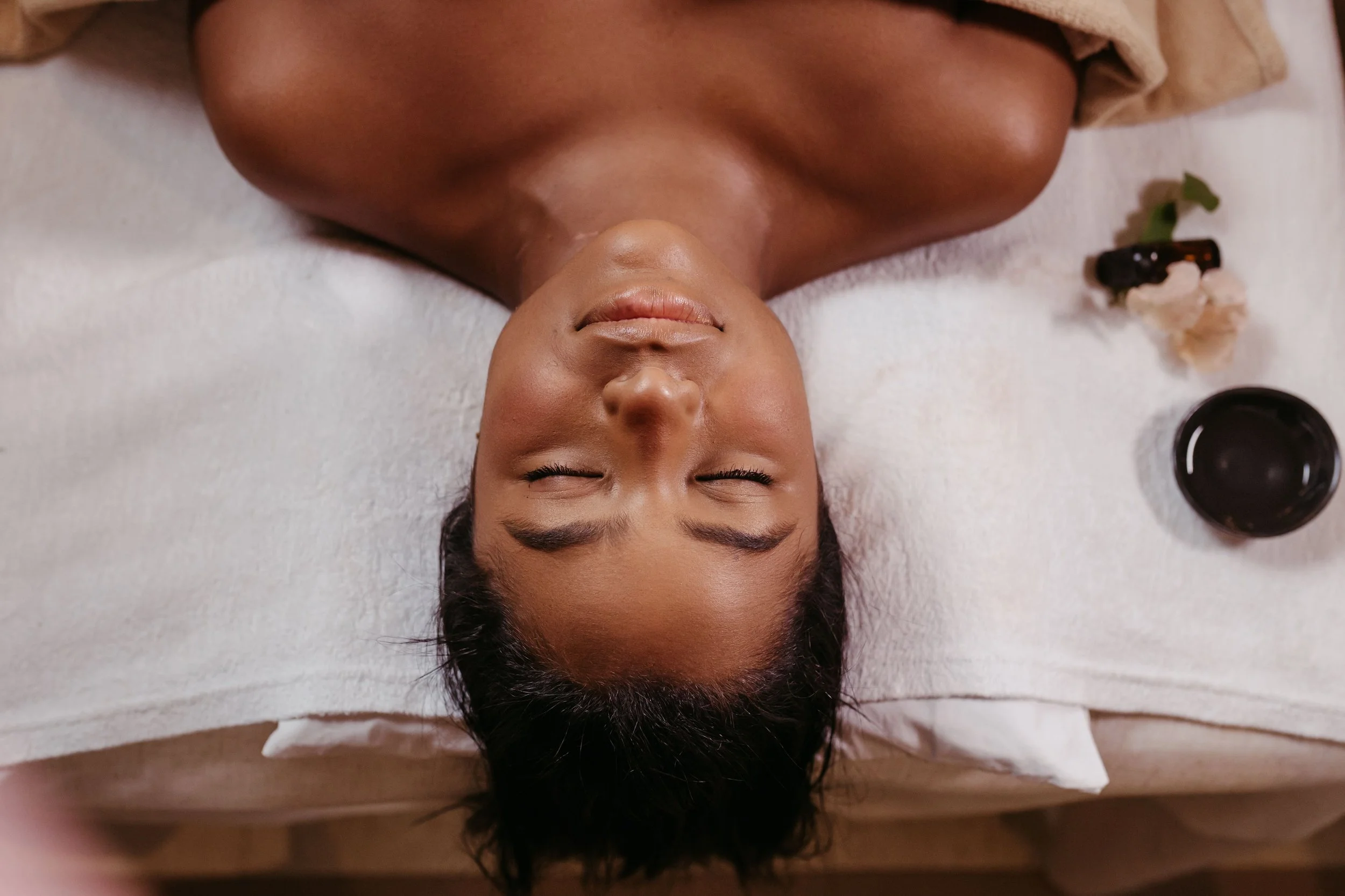 A woman lying on her back with eyes closed, receiving a facial treatment, with skincare products nearby.