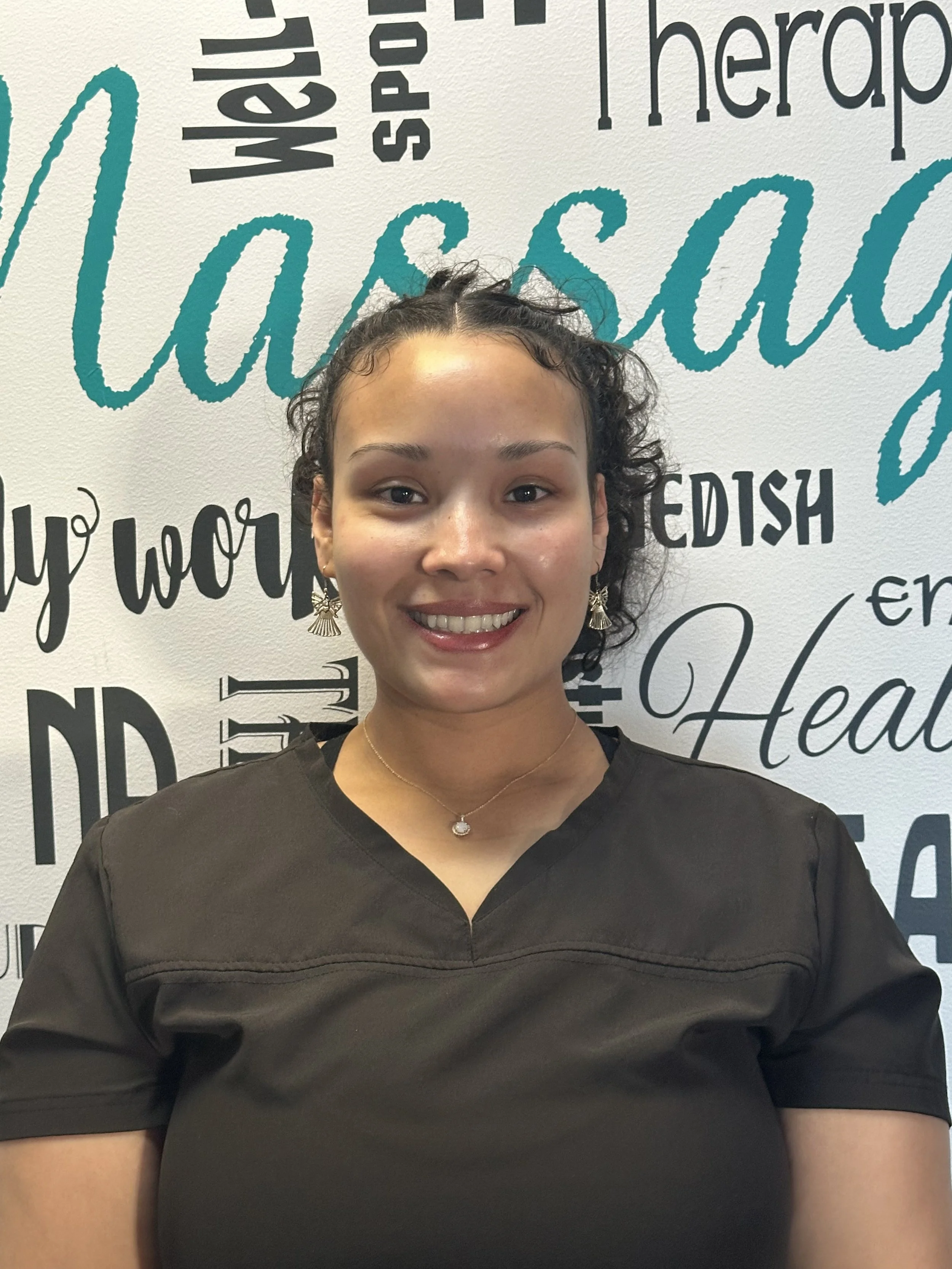 A smiling woman with curly hair wearing earrings, a necklace, and a black shirt, standing in front of a wall with words about wellness, therapy, health, and wellness written in various fonts and colors.