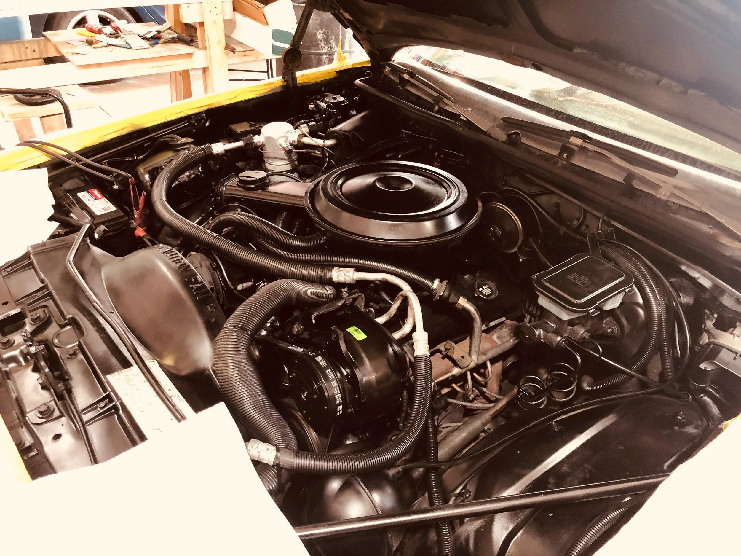 A car engine bay with various hoses, wires, and mechanical components, some of which are covered with a black plastic air filter housing. There is a yellow masking tape along the edge of the open hood and a garage workspace with tools and storage shelves in the background.