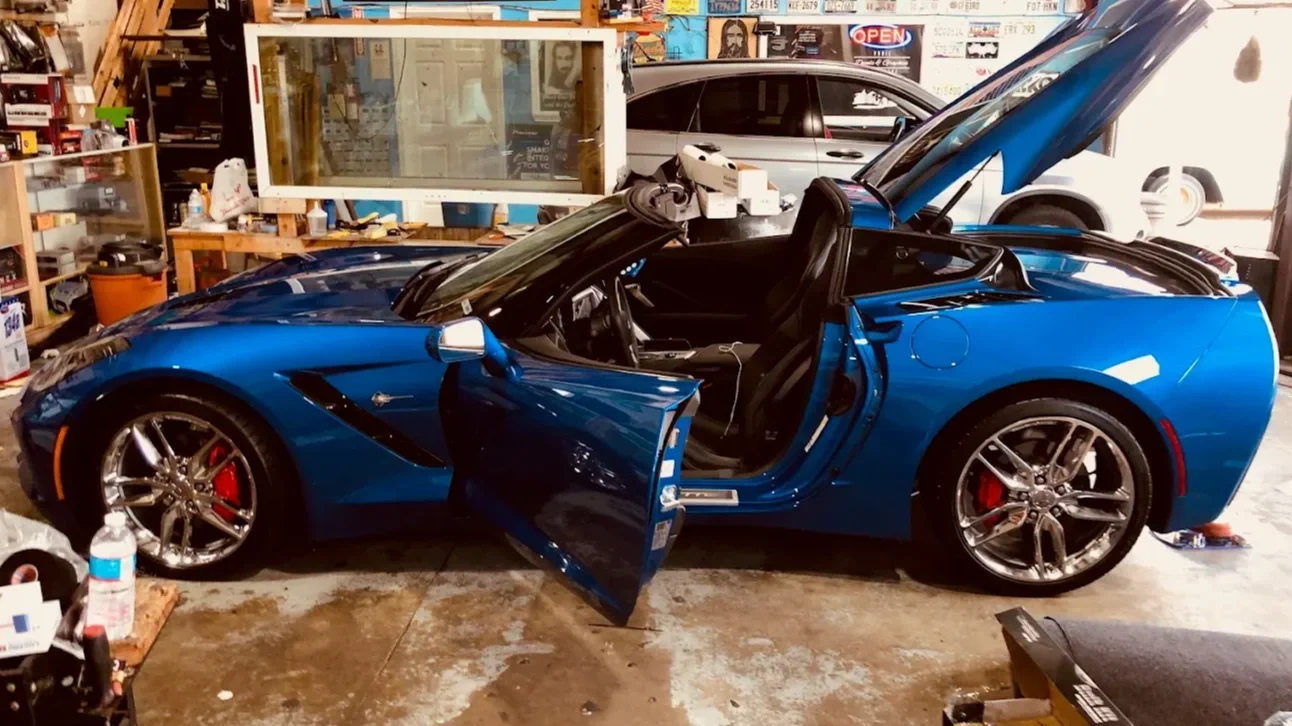 A blue convertible sports car with its doors open and one hood raised, parked inside a cluttered garage.