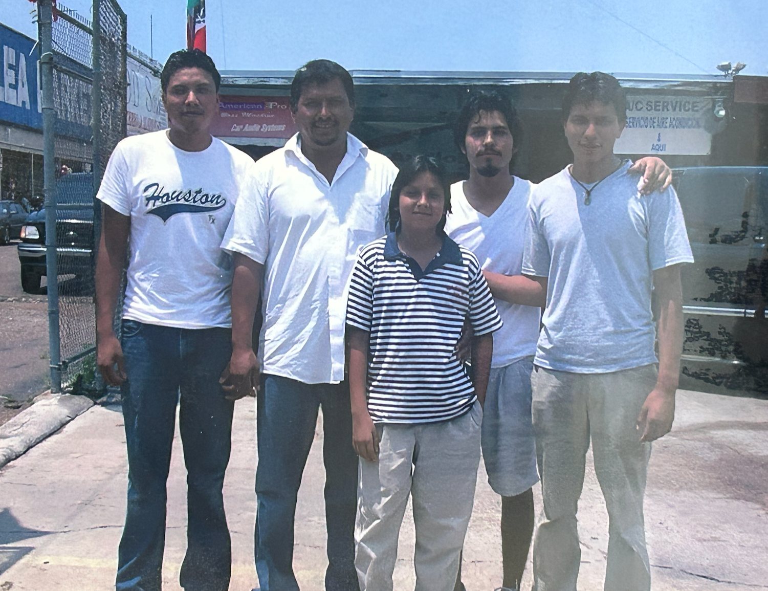 Five people standing outside on a sunny day, posing for a photo. Four men and one young girl are present. The men are wearing casual white shirts, and the girl is in a striped polo shirt with light-colored pants. There are buildings and cars in the background.