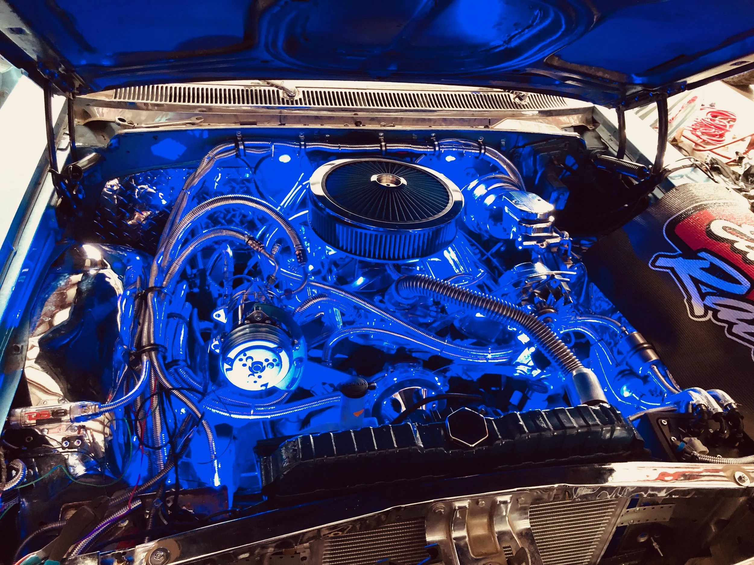 The image shows a car engine bay illuminated with blue light, featuring a large round air filter, various metallic and braided hoses, wires, and mechanical components.