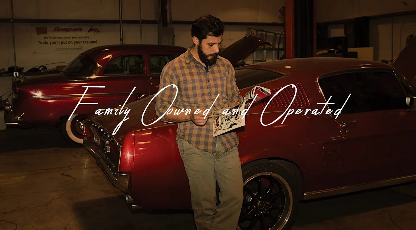 A man with a beard and plaid shirt looking at a brochure while leaning against a red vintage car in a garage with two other classic cars in the background. Overlay text reads, "Family Owned and Operated."
