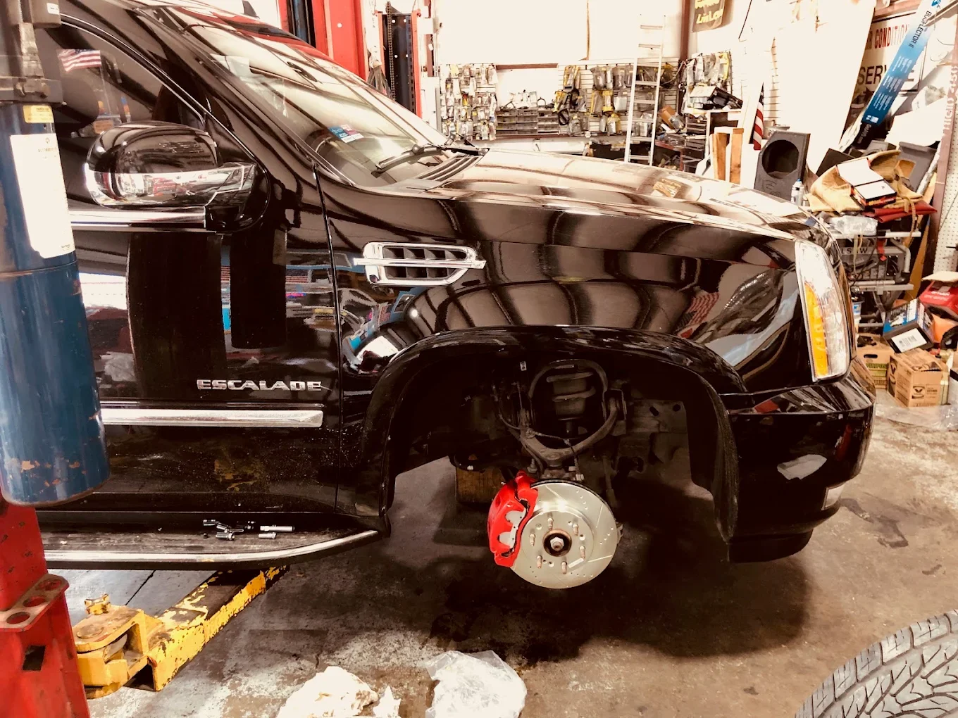 A black Cadillac Escalade SUV in a repair shop with the front wheel and tire removed, exposing the brake caliper and rotor, surrounded by tools and shelves with supplies.