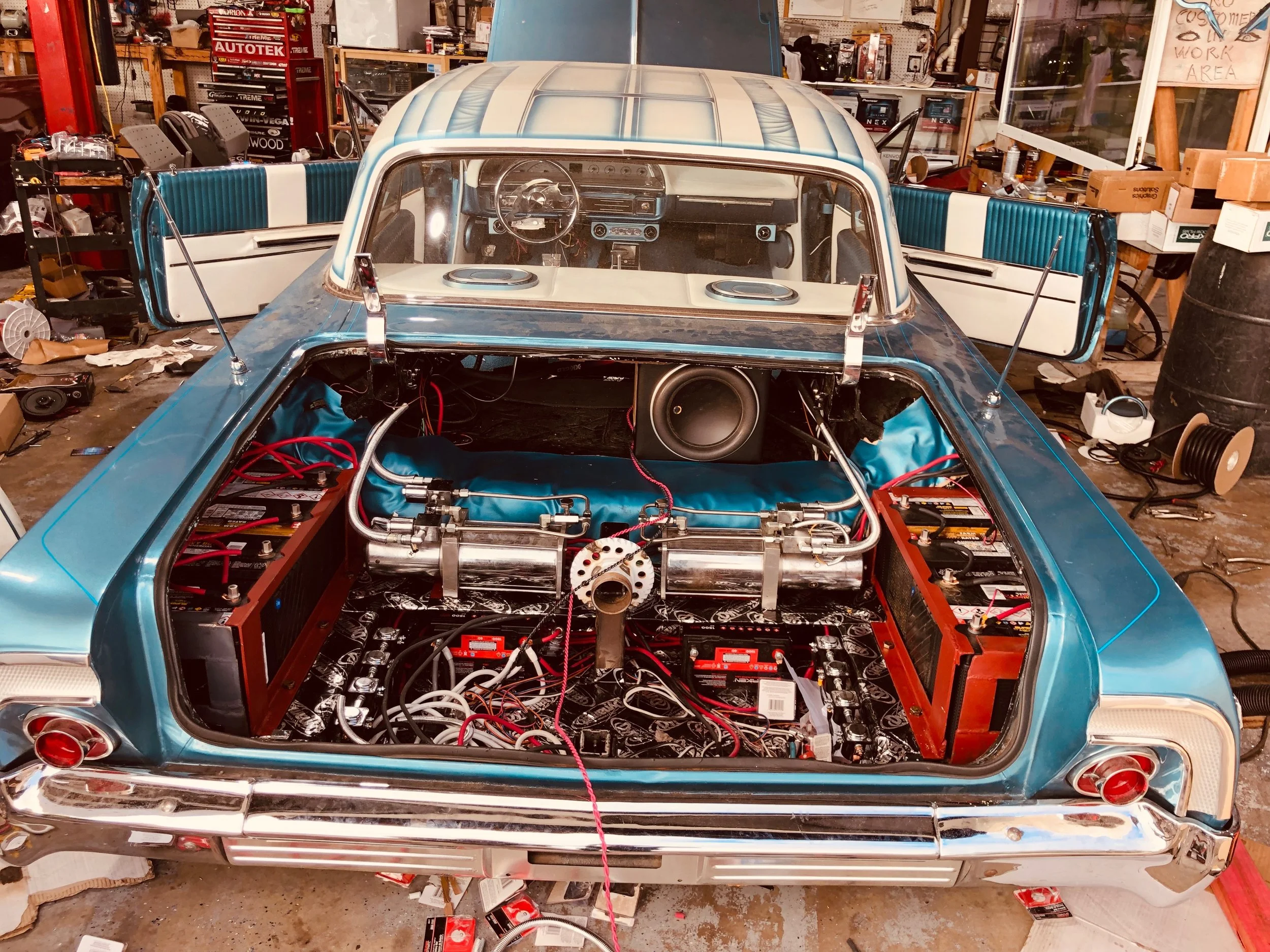 A vintage blue car in a garage, with the rear and trunk open, showing the stripped interior, wiring, and audio system components. The garage has tools, car parts, and clutter surrounding the vehicle.
