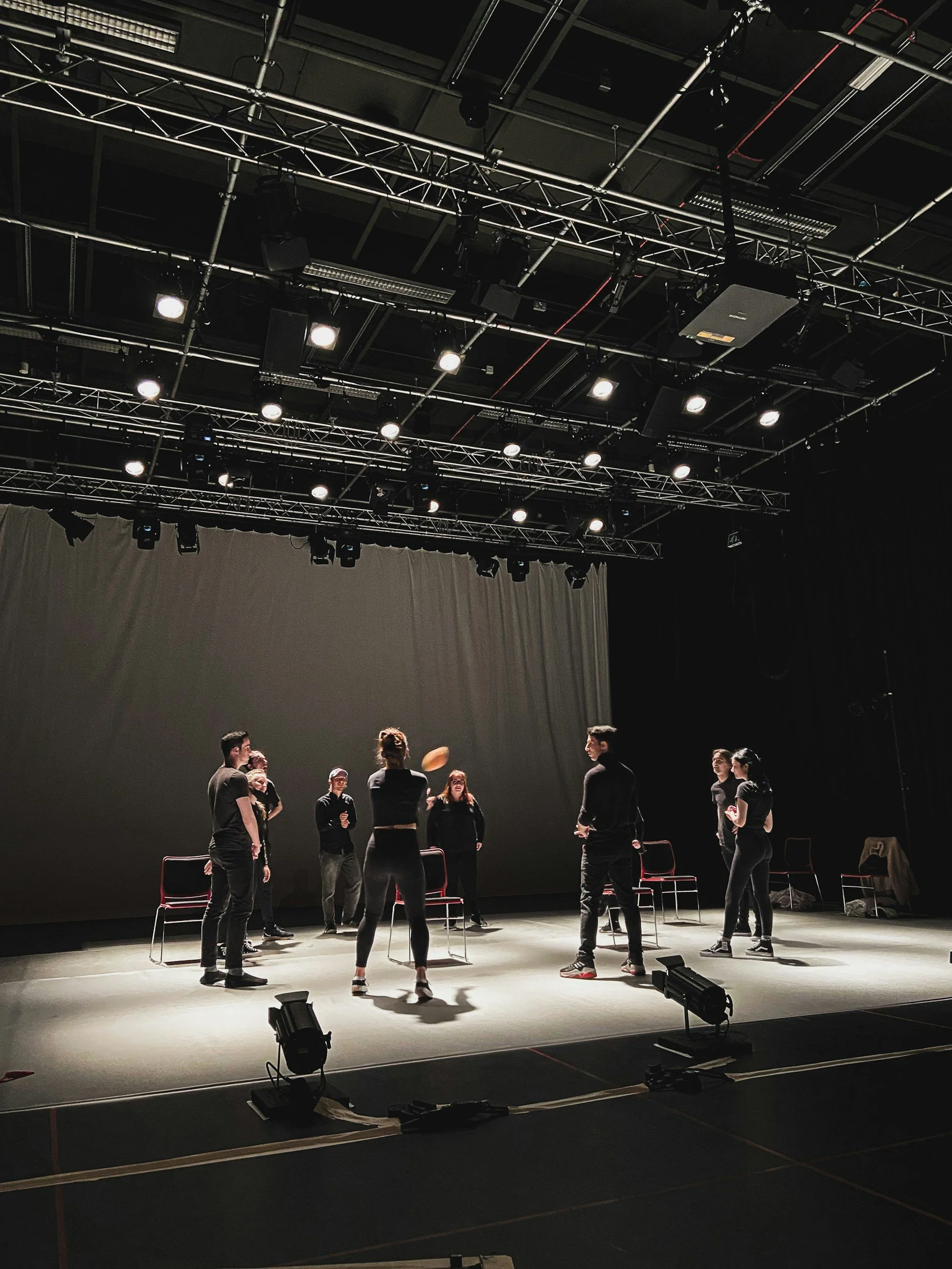 Group of people rehearsing or performing on a stage with chairs, lights, and a curtain backdrop in a theater or studio setting.