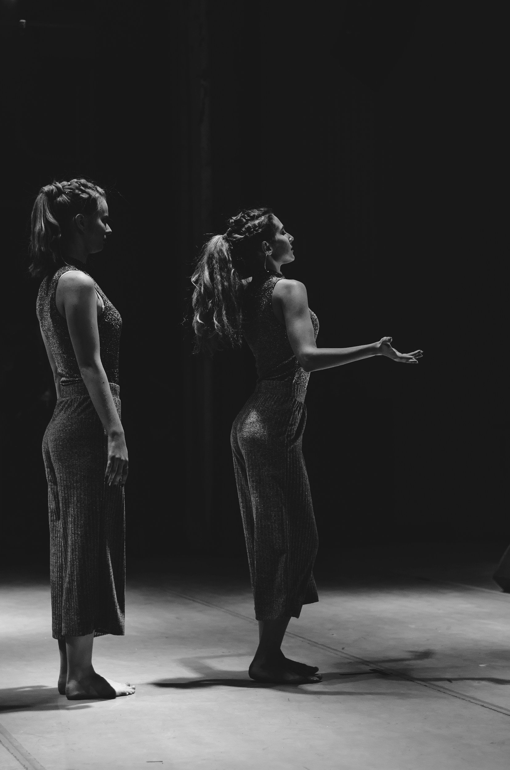 Two women standing barefoot on a stage, facing left, with one woman extending her hand outward, dressed in matching sleeveless tops and wide-legged pants, in what appears to be a dance or performance setting.