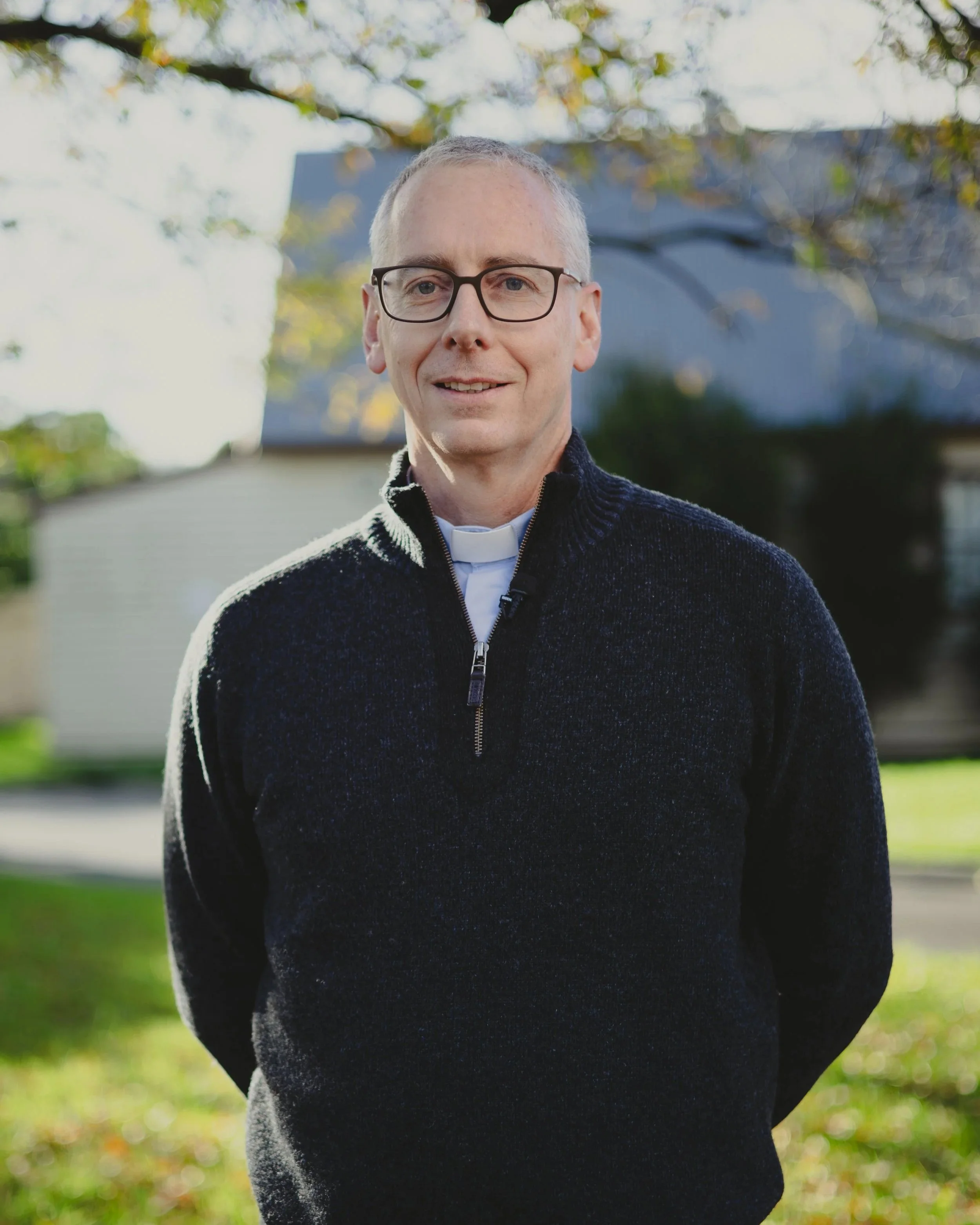 A middle-aged man with gray hair, wearing glasses, a clerical collar, and a dark zip-up sweater, standing outdoors with trees and a house in the background.
