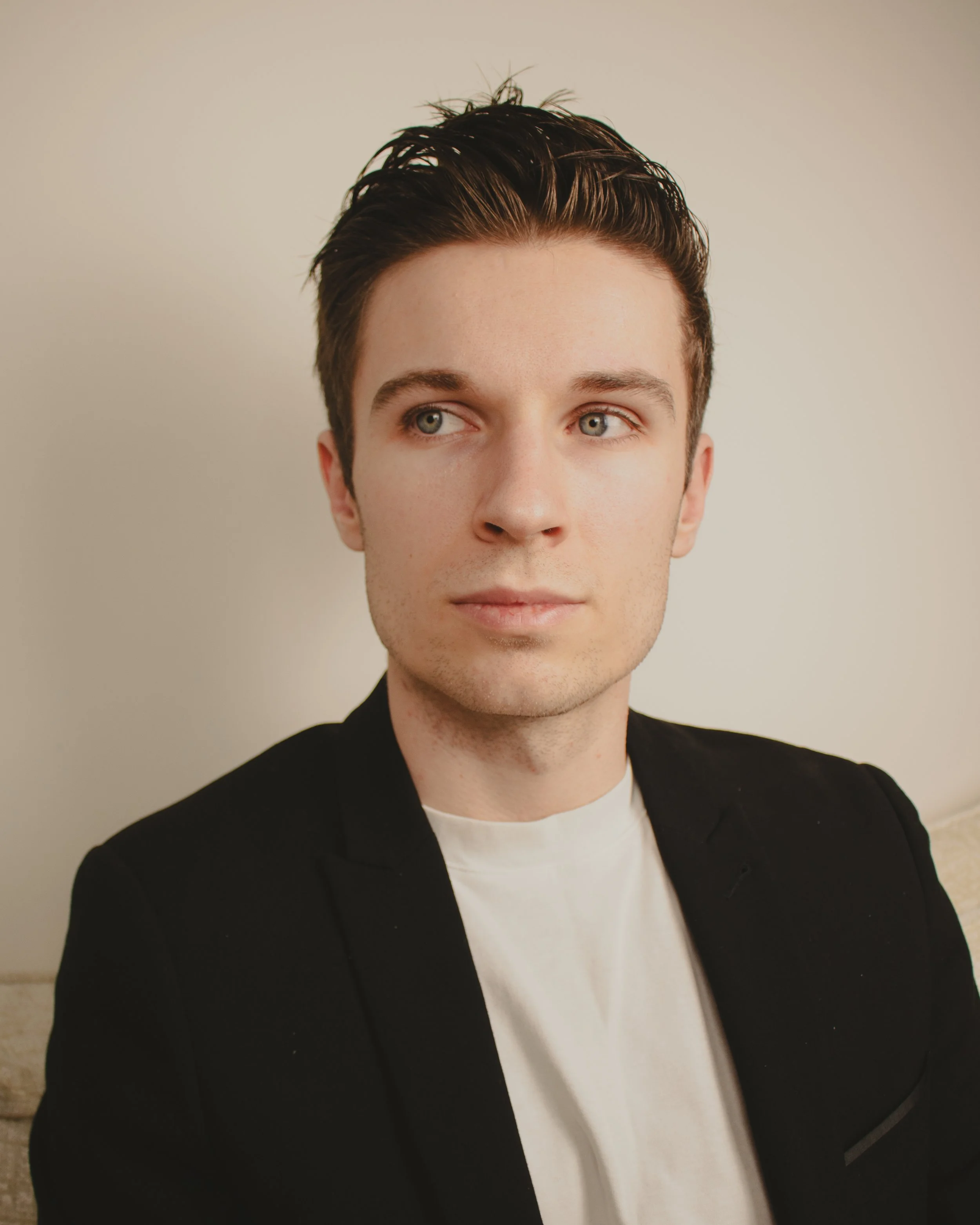 A young man with dark brown hair styled back, light skin, wearing a black blazer over a white shirt, looking slightly to the side, with a neutral expression, standing against a plain light-colored wall.