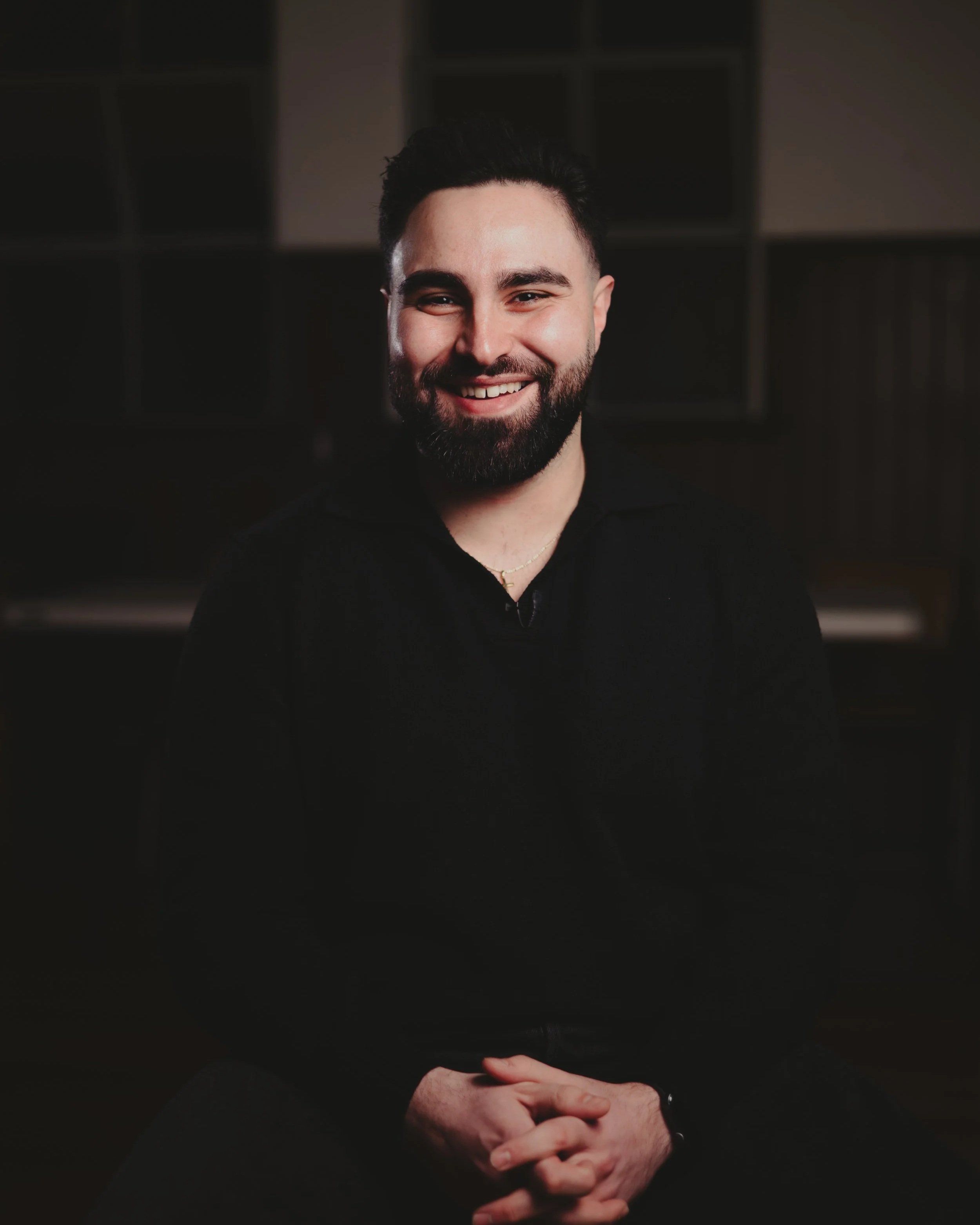 A man with a beard smiling, dressed in black, with hands clasped in front of him, standing against a dark background with geometric patterns.