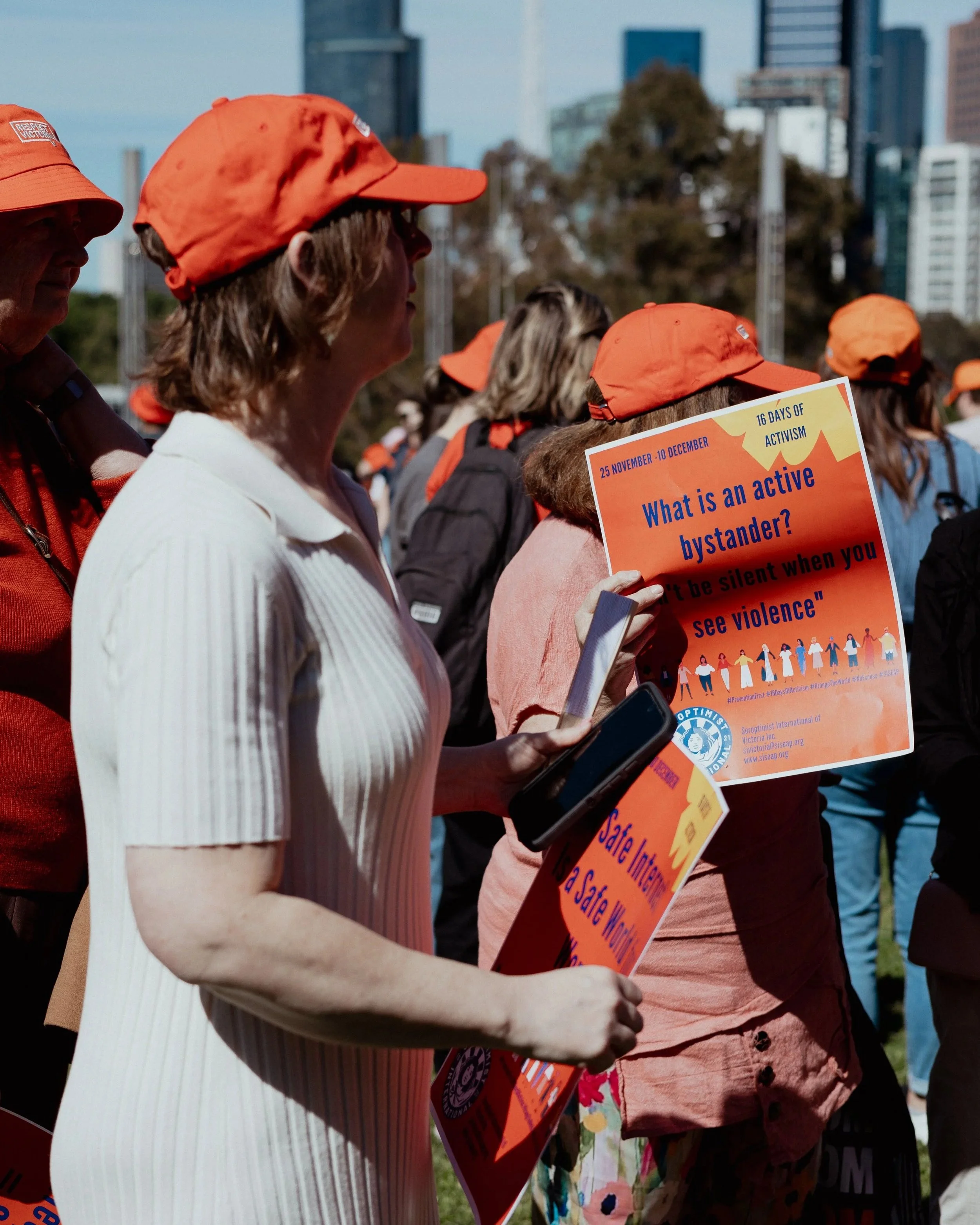 People gathered outdoors wearing orange hats, participating in a protest or rally, with one holding a sign about active bystanders. Prevent Violence Against Women Walk.