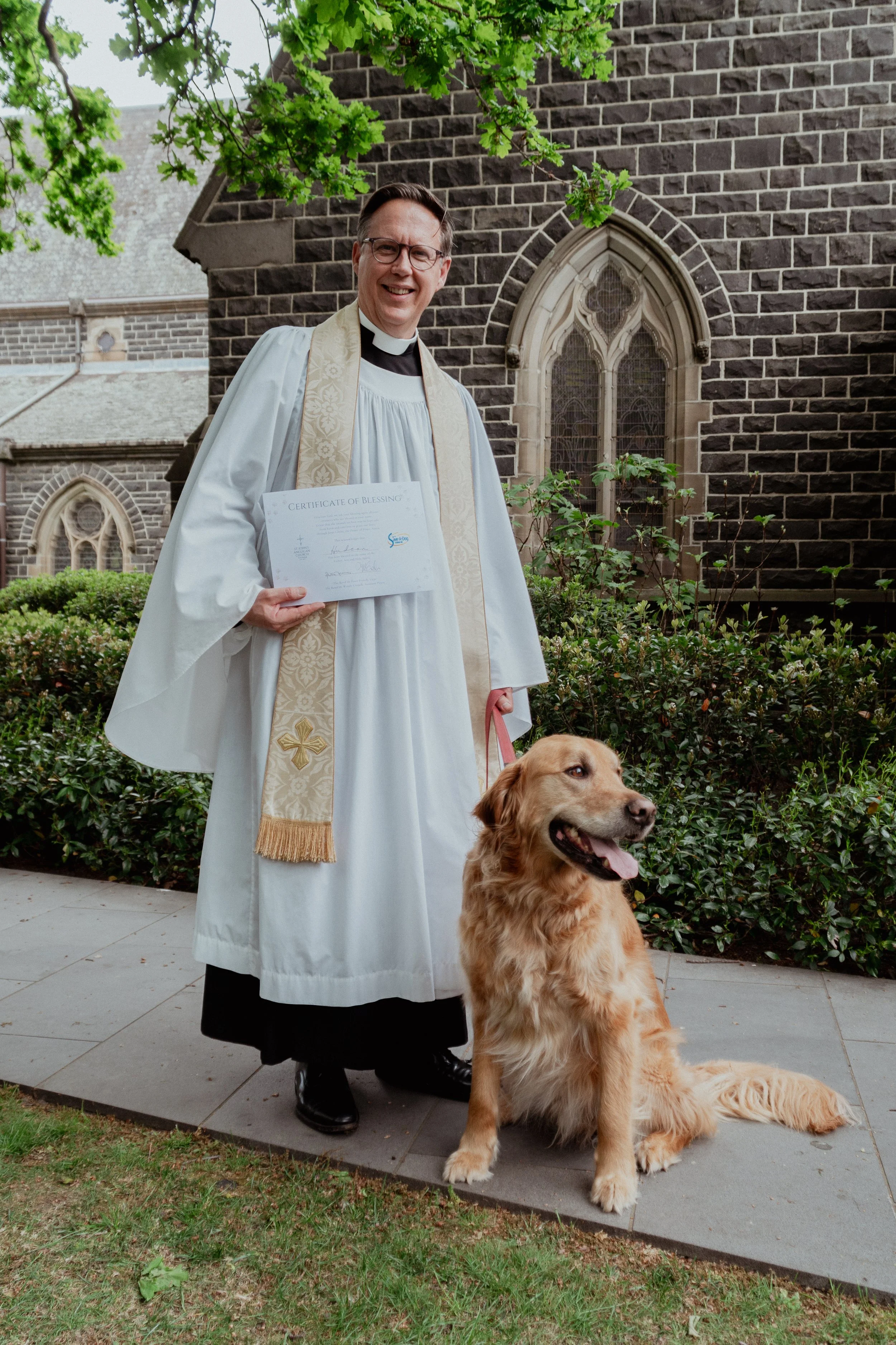 A smiling male priest in a white robe holding a certificate of blessing, standing outside a brick church with green trees, and sitting next to a large golden retriever dog.