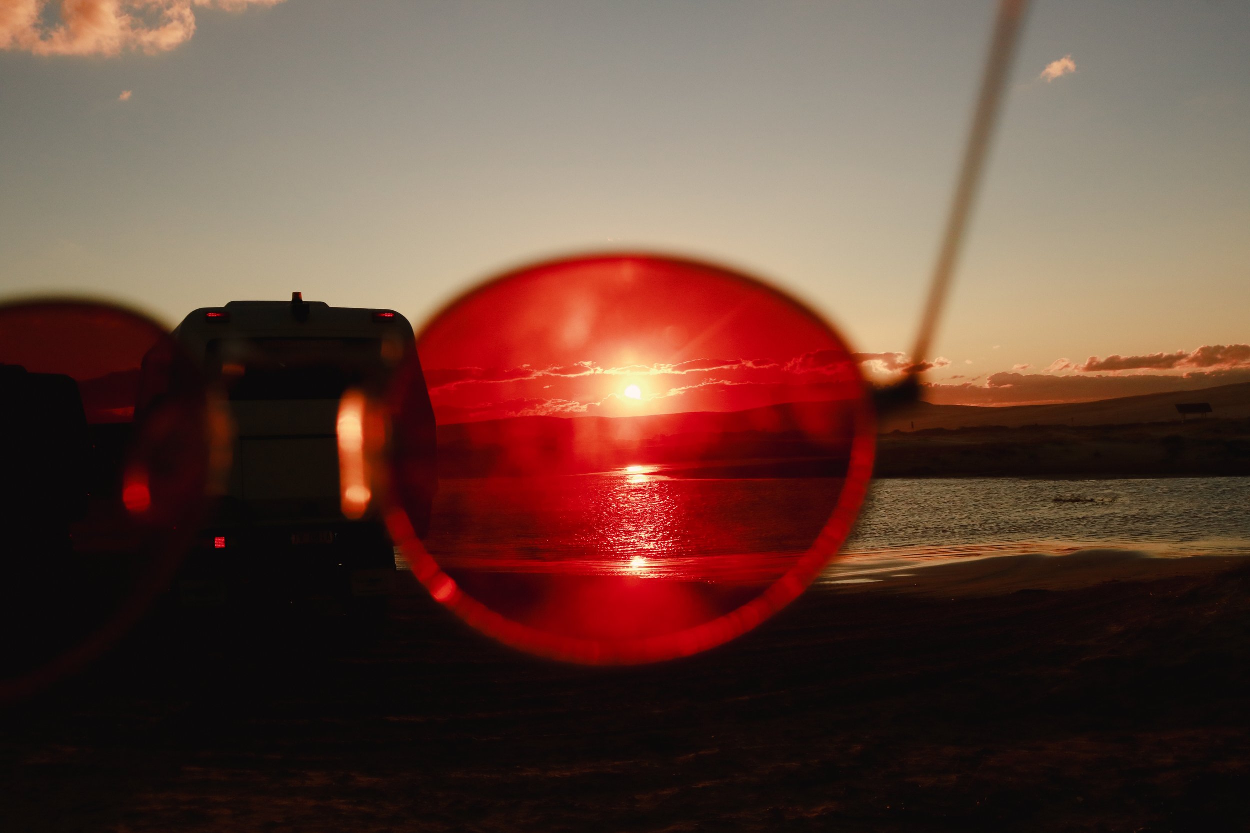 Sunset over water seen through the red-tinted lenses of sunglasses with a camera silhouette in the foreground.